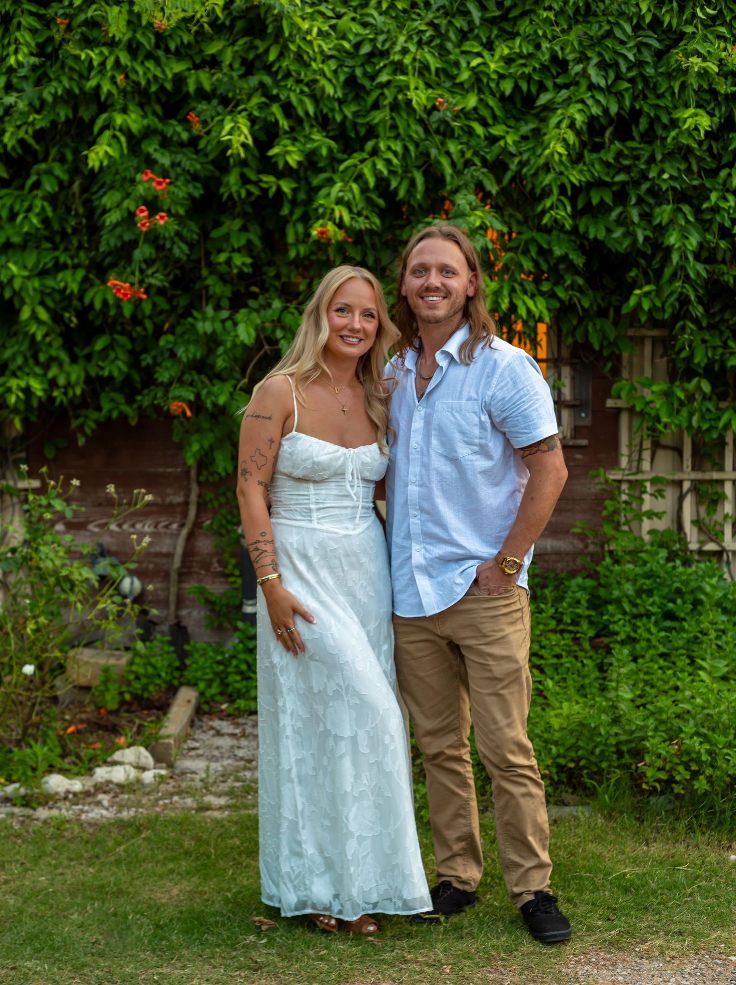 Couple smiling together in a lush garden, woman in white dress and man in light blue shirt and khaki pants.