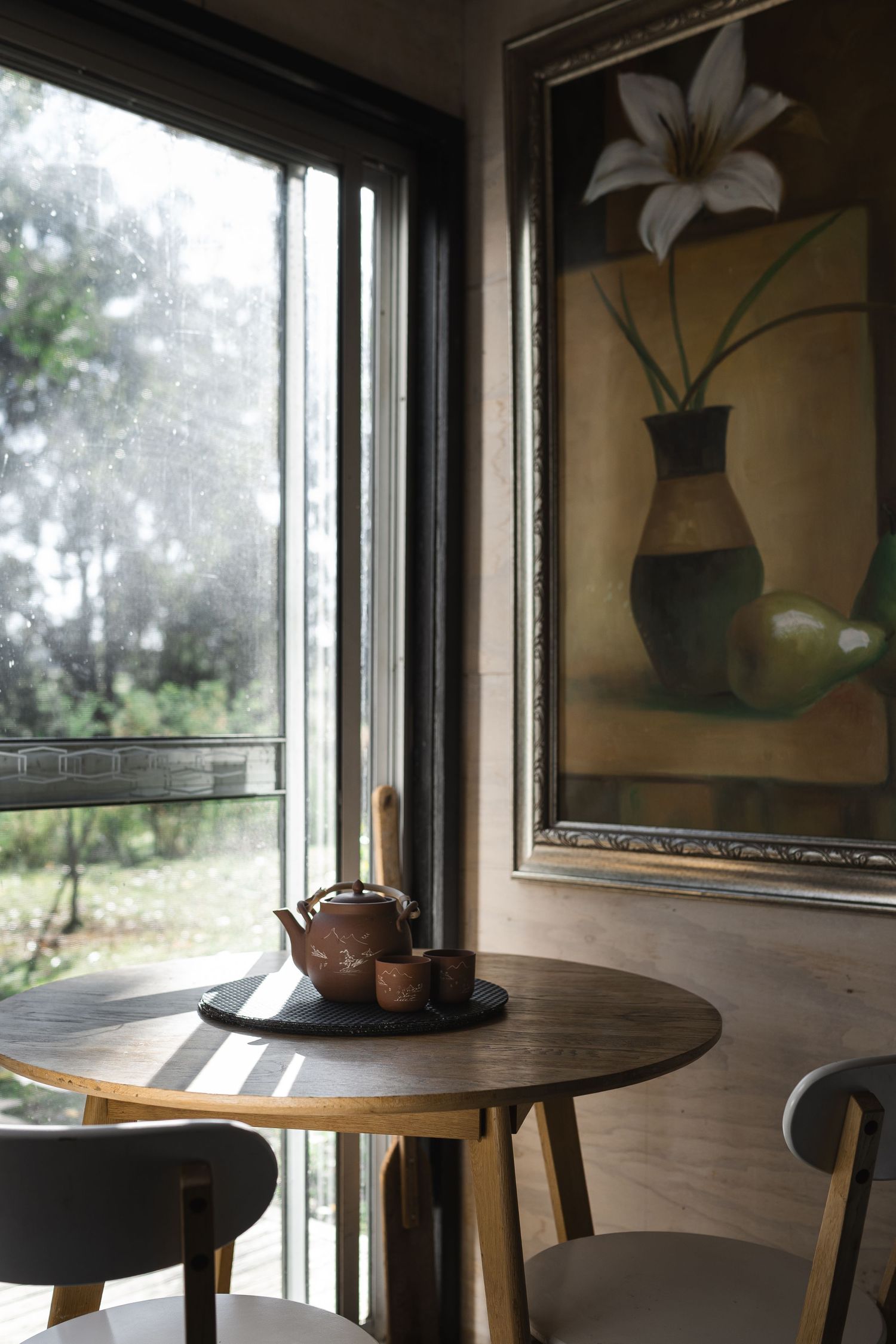 Cozy cafe corner with round wooden table, clay teapot set, and framed floral painting beside a rain-streaked window.
Cedar & Stone