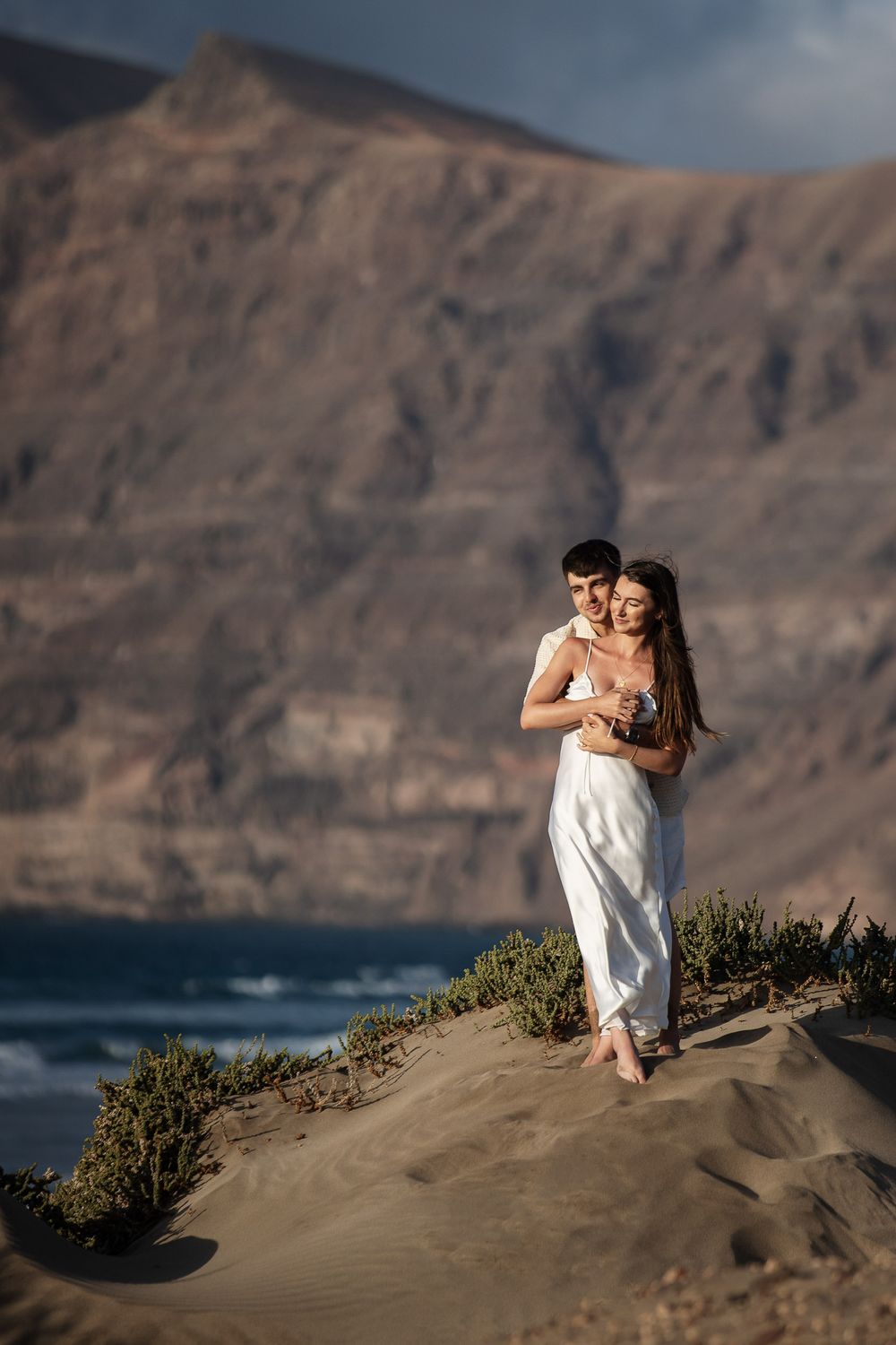 Couple embracing on sandy dune with ocean and dramatic cliffs backdrop, woman in white dress, romantic outdoor photo.