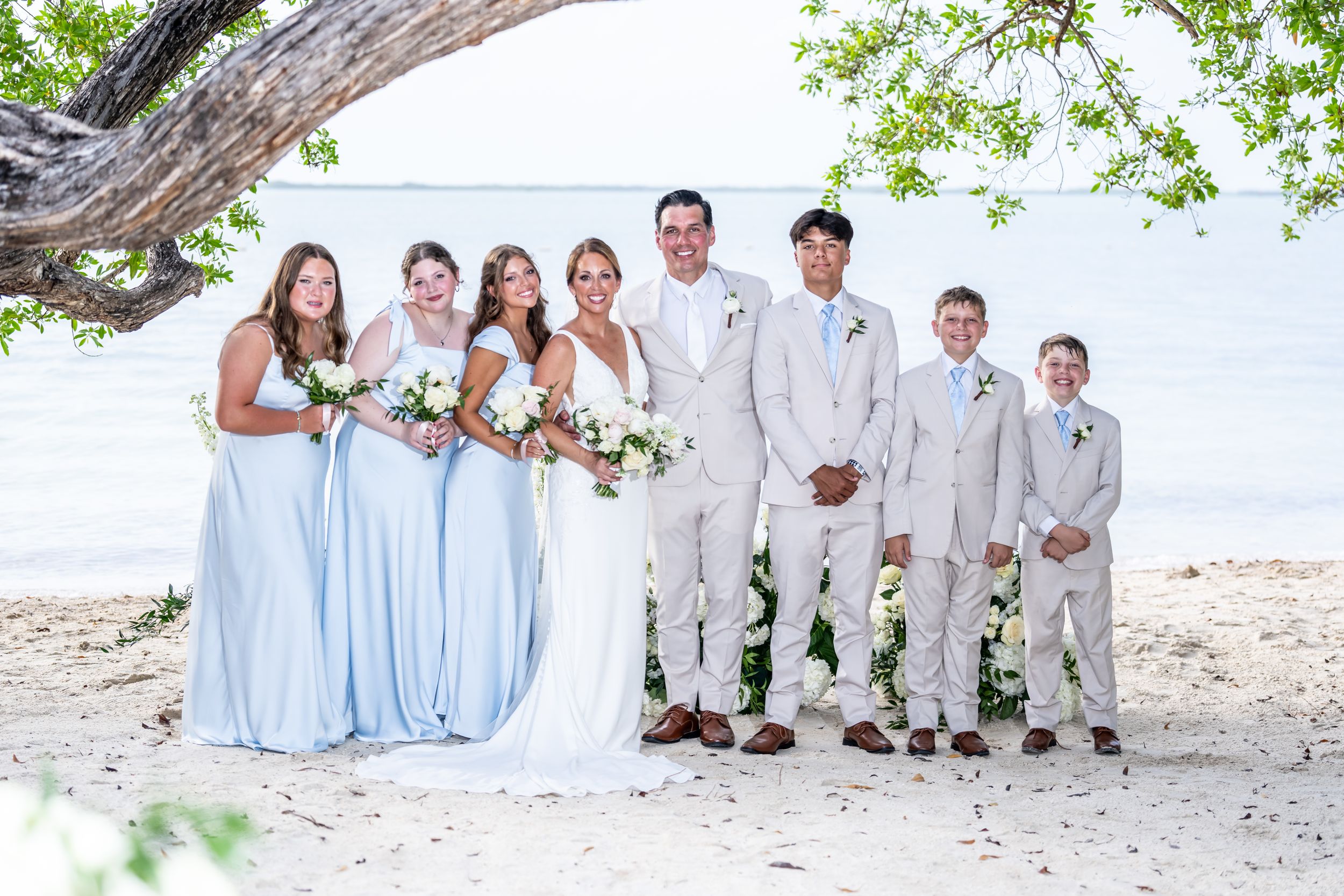 Wedding party posing on a sandy beach, bride in white gown, bridesmaids in light blue dresses, groomsmen in beige suits.