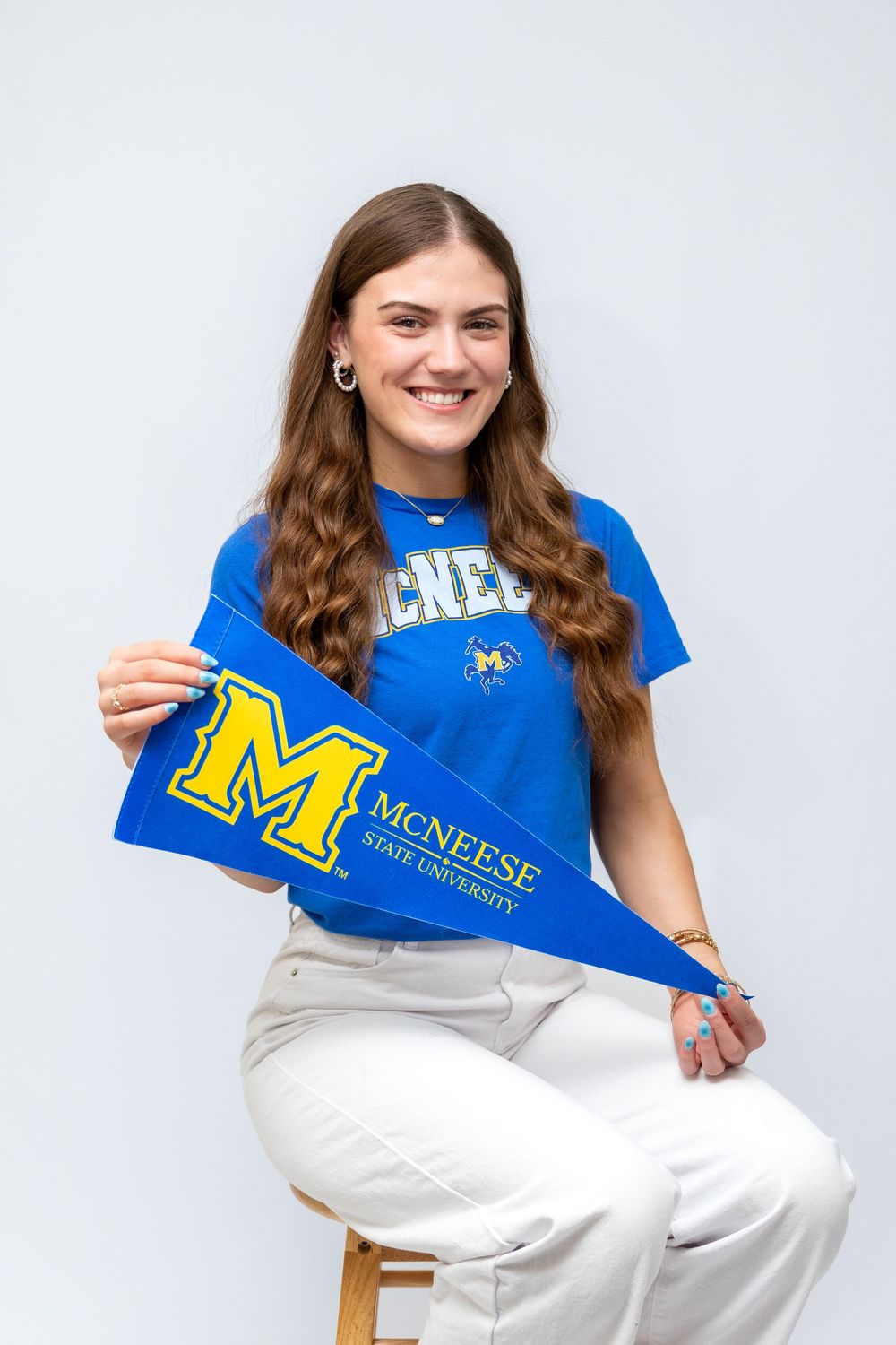 Smiling young woman holding McNeese State University pennant, wearing blue McNeese t-shirt, seated against white background.