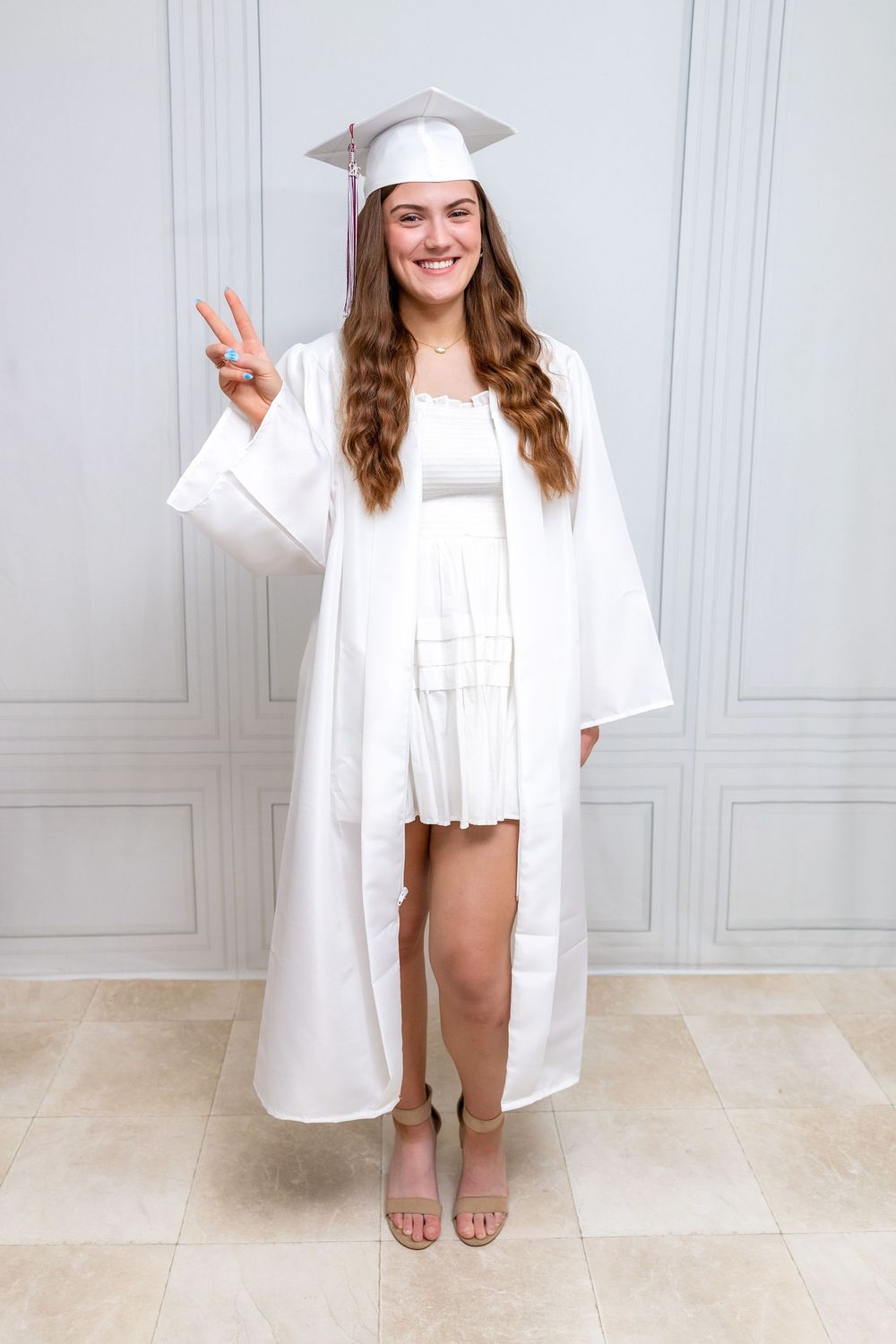 Smiling young woman in white graduation cap and gown making a peace sign, posing for a full-length portrait.