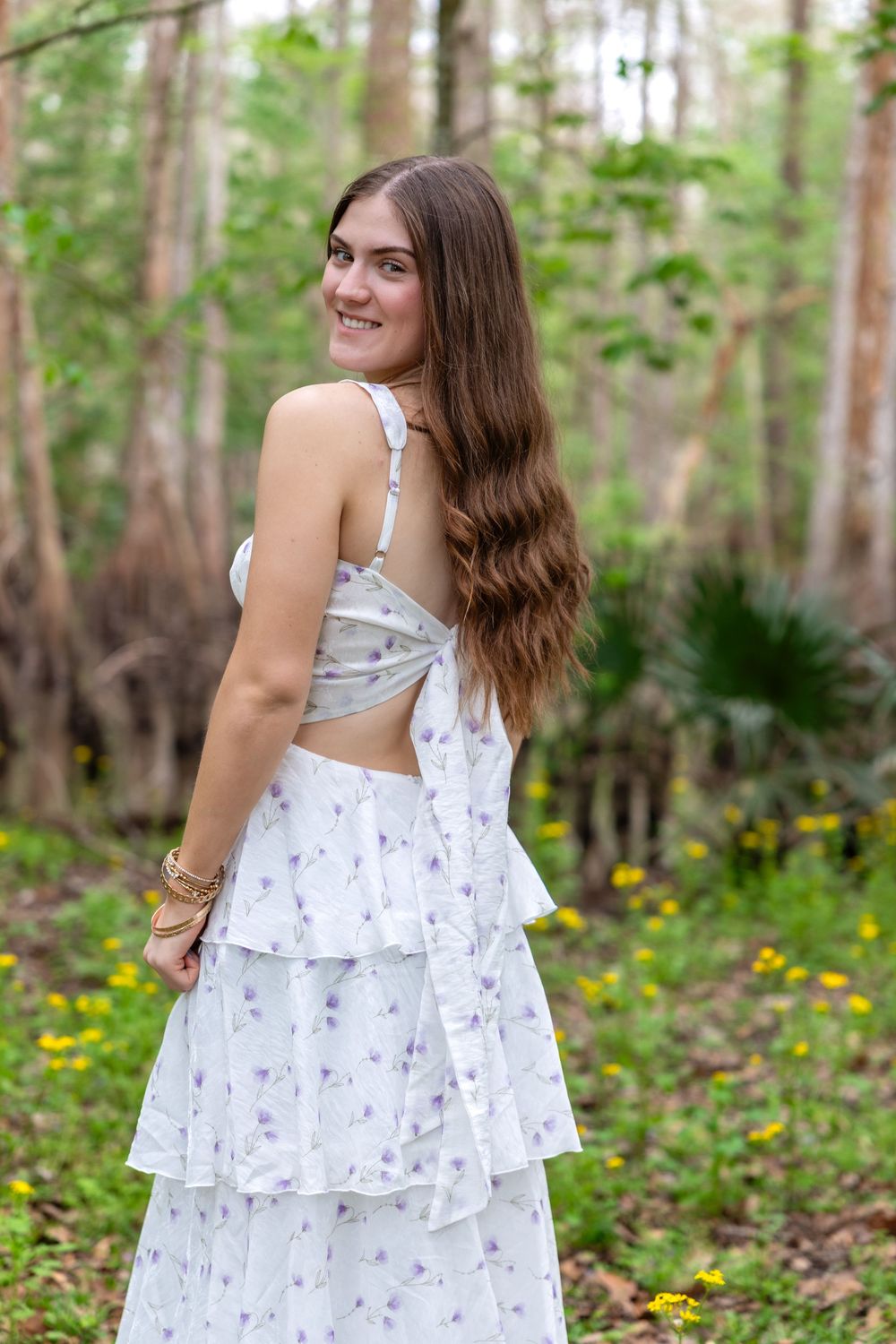 Young woman in white floral tiered dress smiling over shoulder in lush green forest setting.