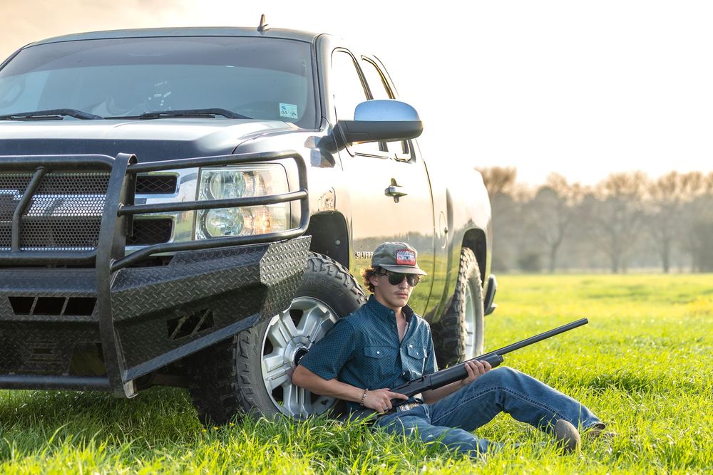 Man sitting in grass leaning against a truck holding a rifle on a sunny day outdoors.