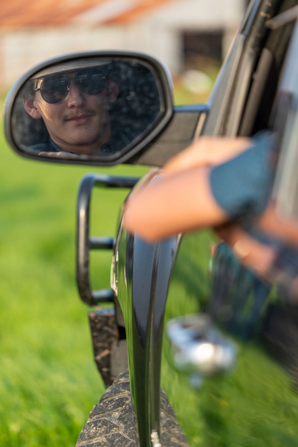Person wearing sunglasses reflected in a car side mirror, leaning out of a vehicle window on a sunny day.