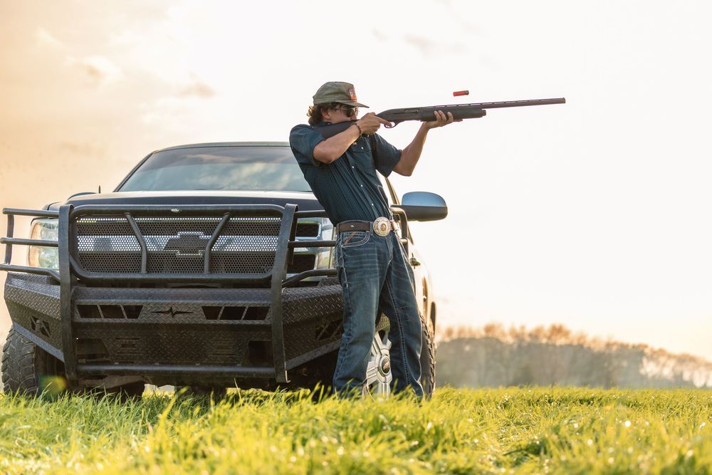 Man aiming a shotgun beside a Chevrolet truck in a rural field at golden hour.