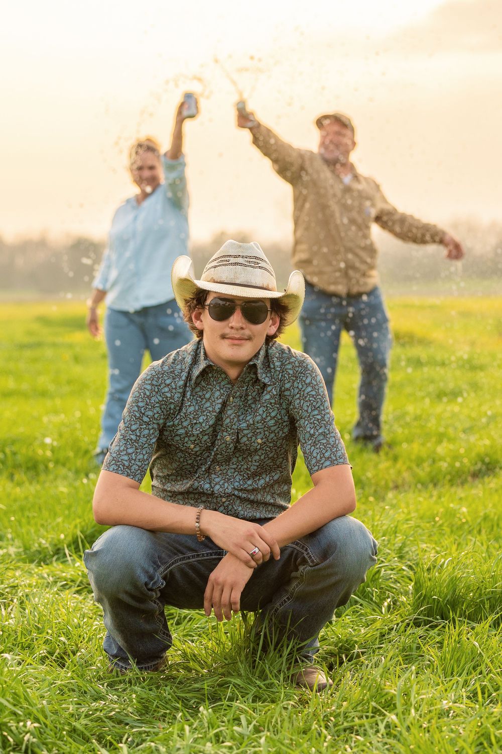 Young man in cowboy hat and sunglasses crouches in green field while couple celebrates with sparklers behind him.