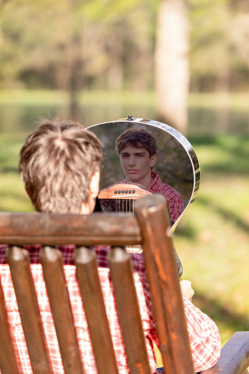 Young man with guitar reflected in instrument's shiny body while sitting in wooden chair outdoors in sunny park.