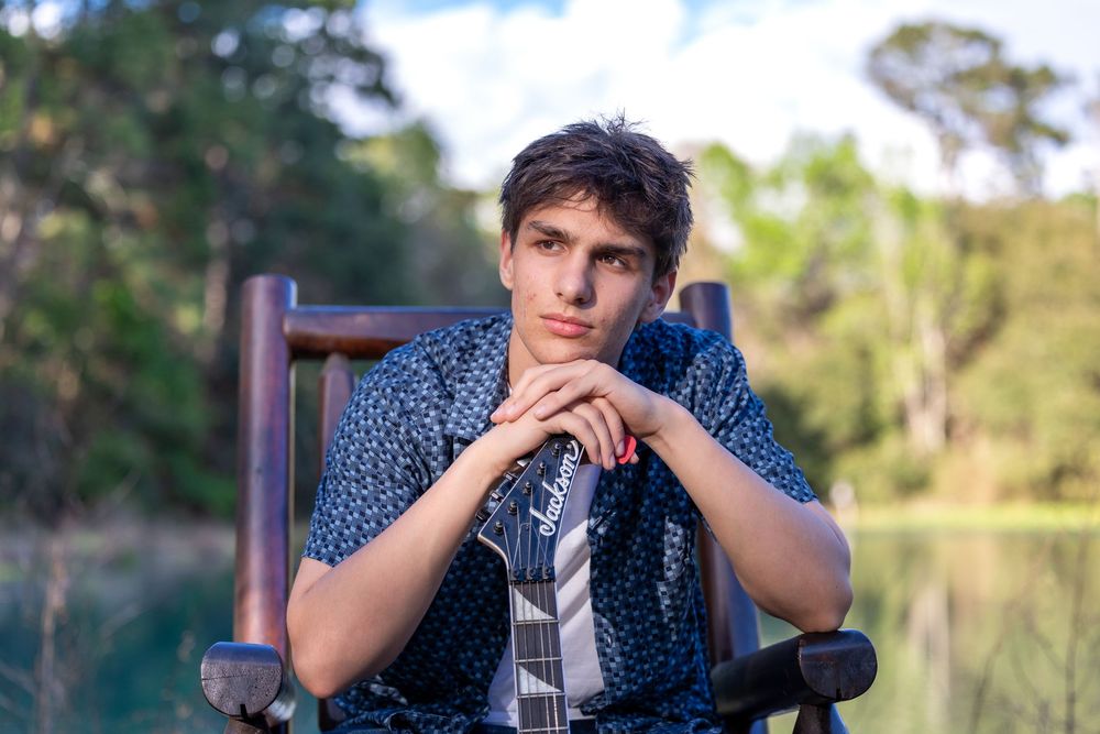 Young man in blue patterned shirt sitting in wooden chair outdoors near a serene lake surrounded by trees.