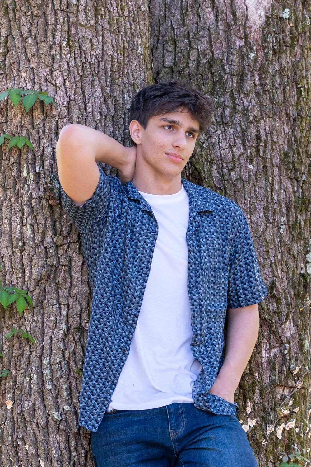 Young man in blue patterned shirt and jeans leaning against large tree trunk outdoors.