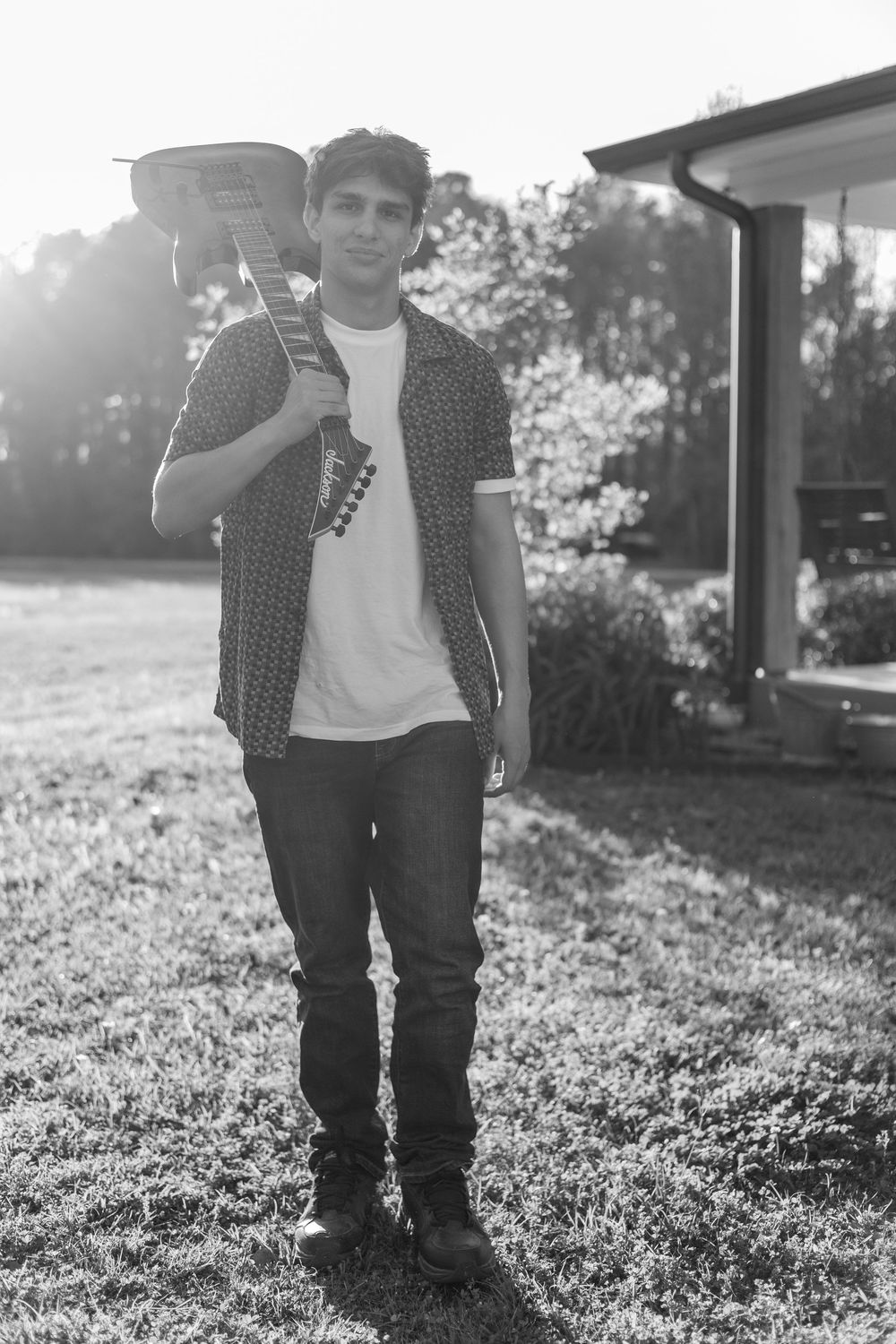 Young man holding a guitar over his shoulder standing on grass outdoors in a black and white photo.