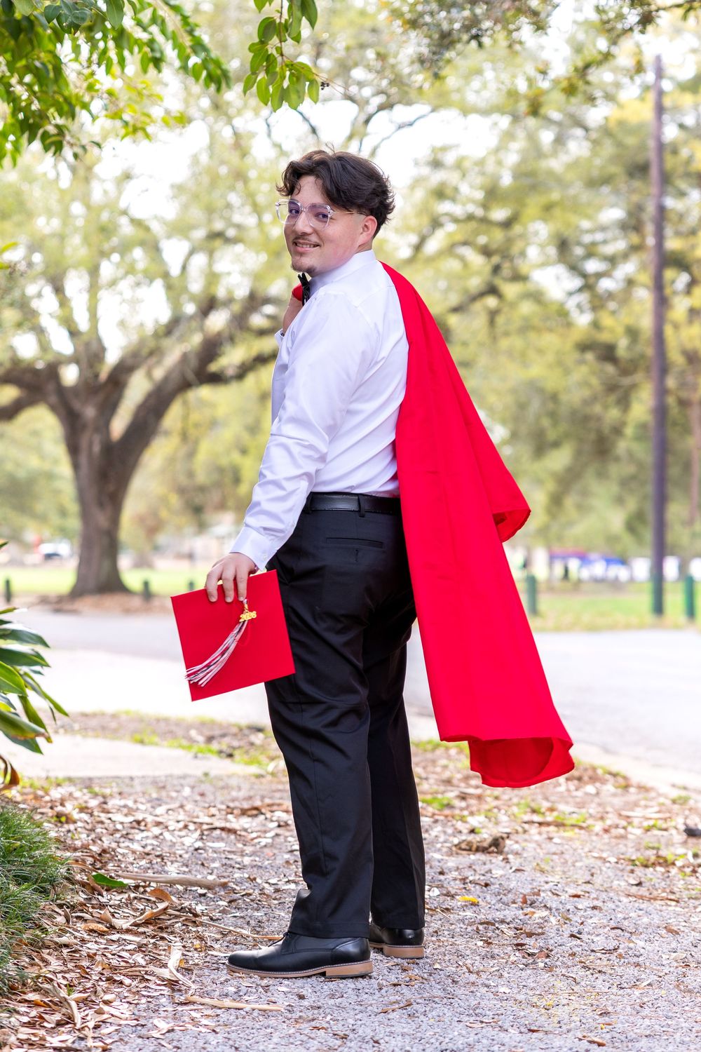 Young man in white shirt and red graduation gown holds cap outdoors among trees, smiling over shoulder.