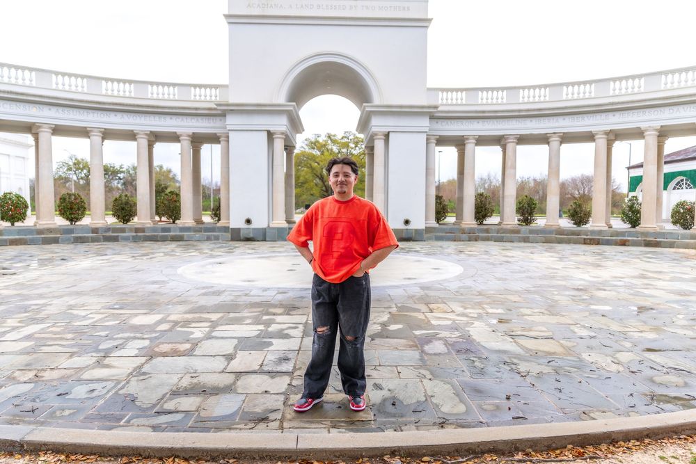 Person in red shirt standing in front of elegant white colonnade archway on stone plaza outdoors.