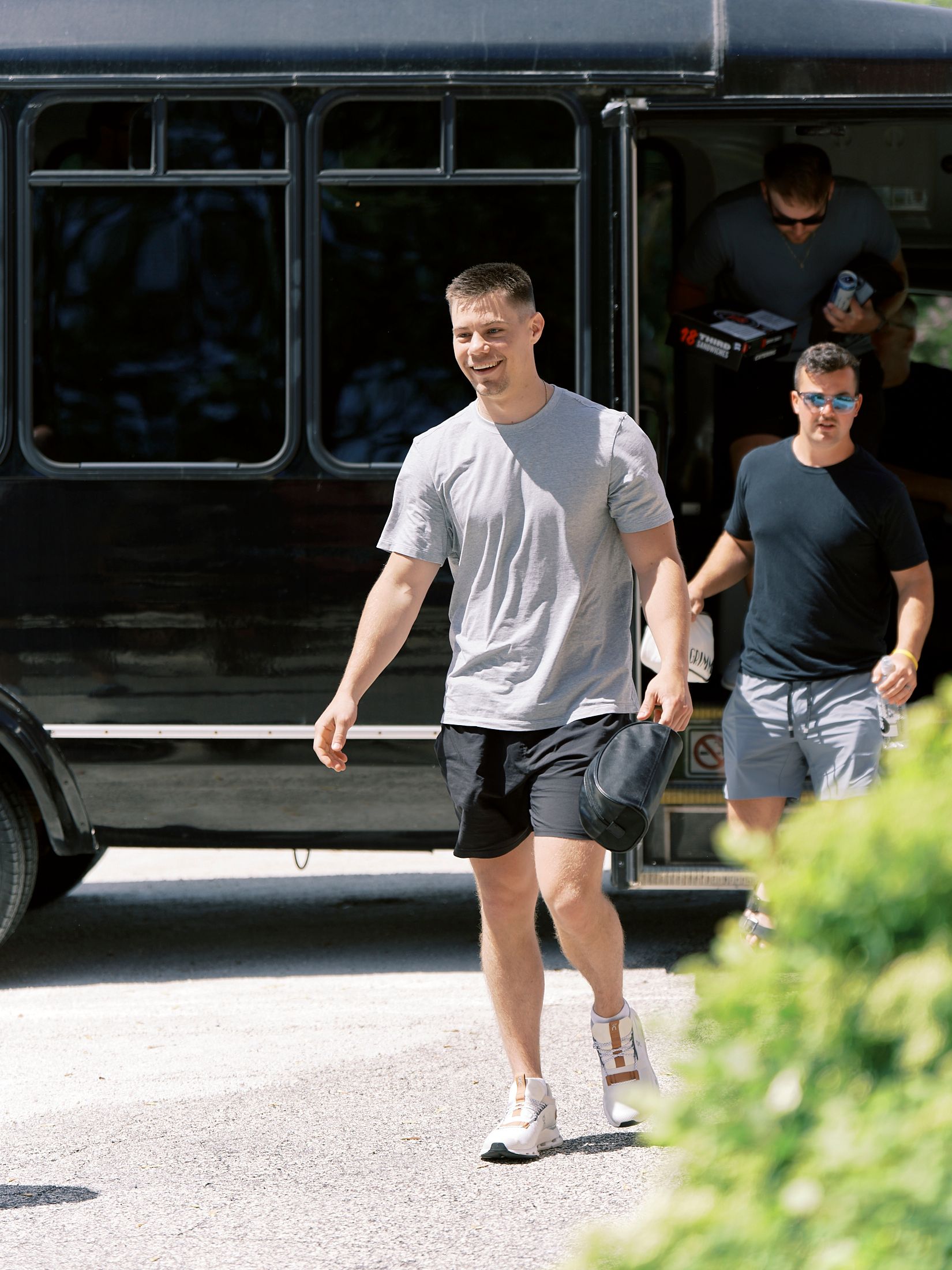 Two men in casual athletic wear walking away from a black bus on a sunny day, smiling outdoors.