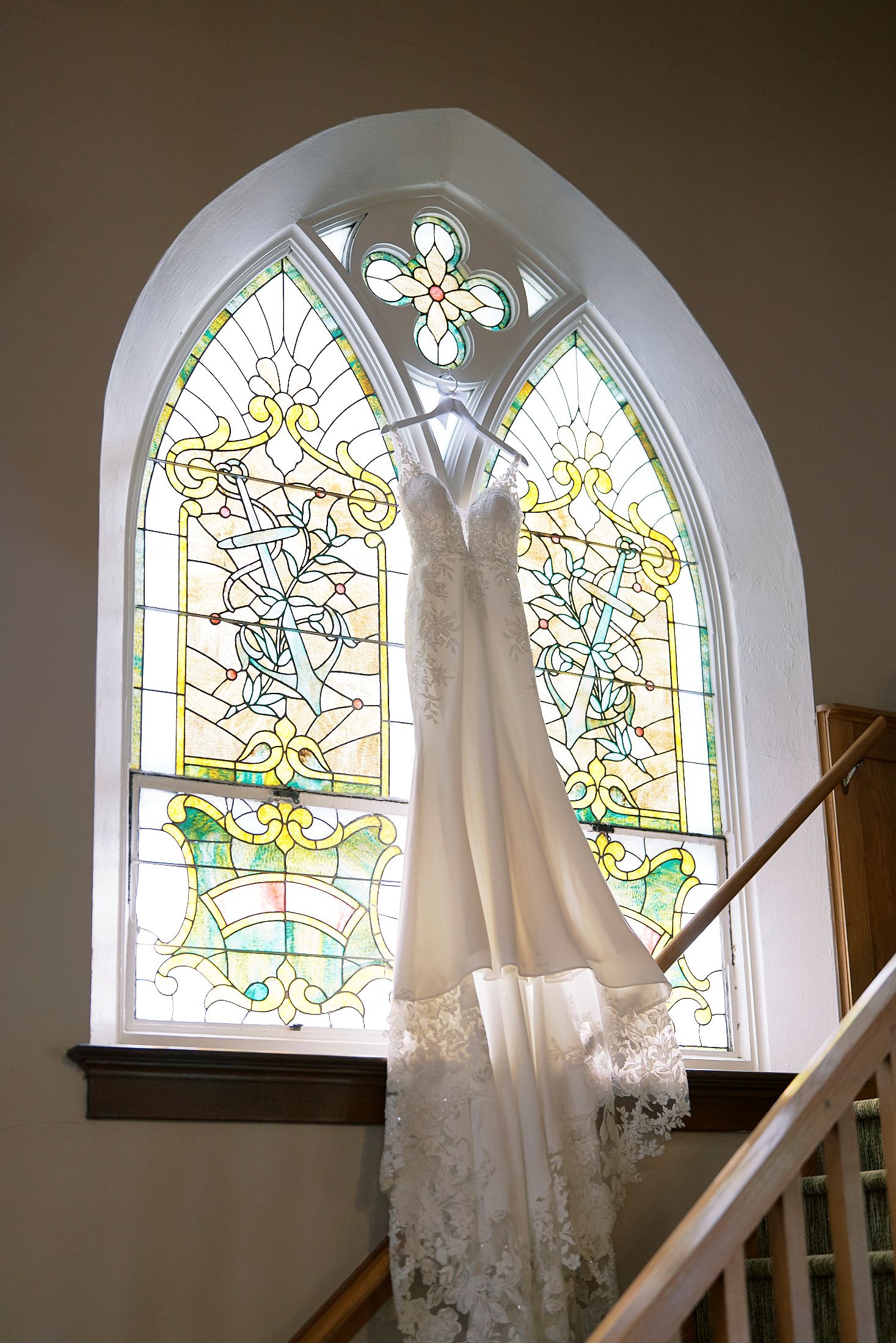 A white lace wedding dress hanging in front of a colorful stained glass arched church window above a wooden staircase.