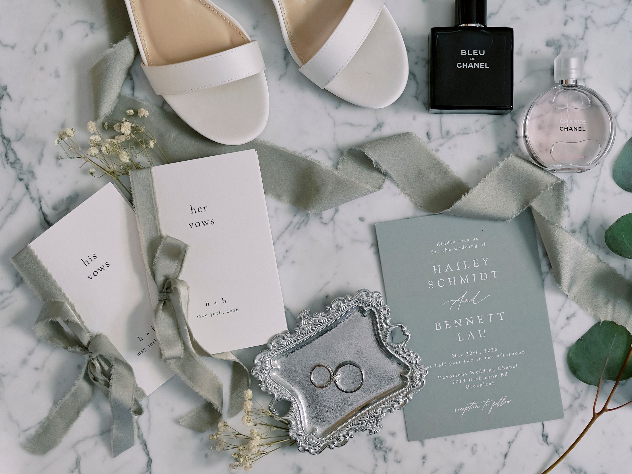 Elegant wedding flat lay with sage green invitations, wedding rings, white heels, and Chanel perfume on marble.