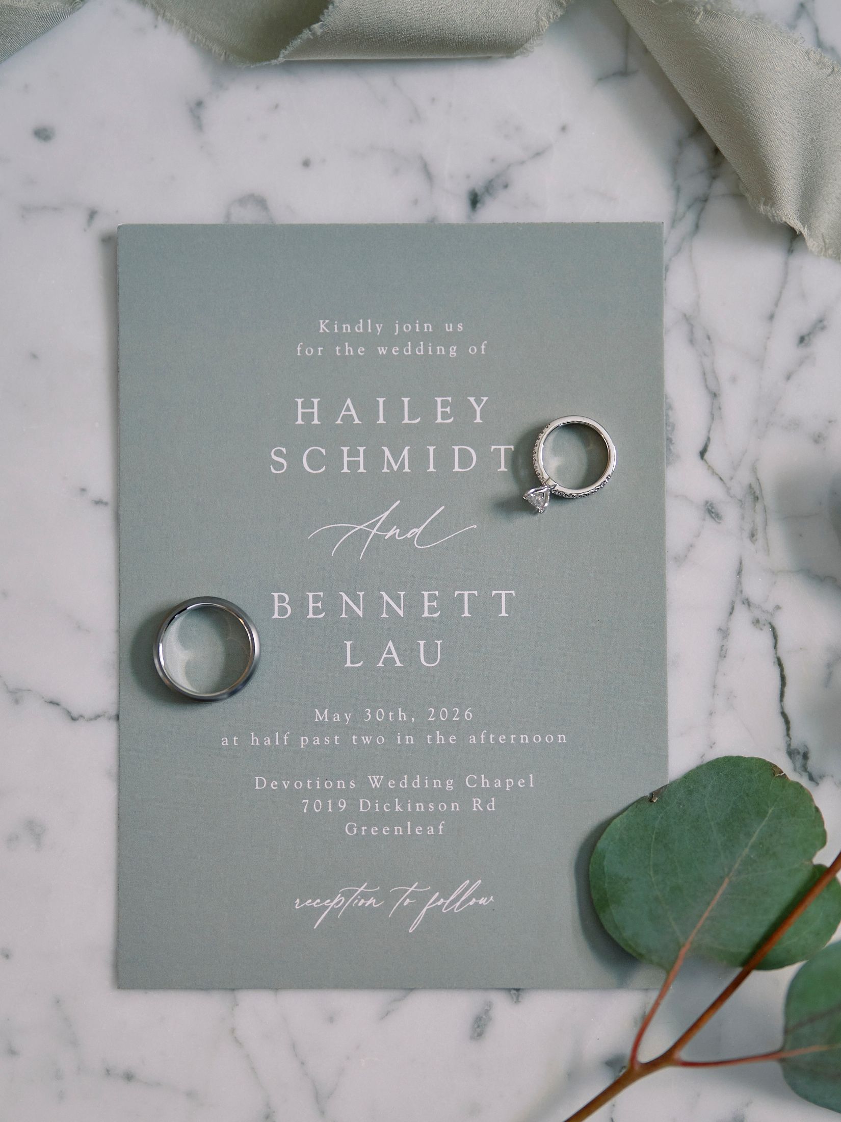 Elegant sage green wedding invitation for Hailey Schmidt and Bennett Lau with gold rings on marble surface.