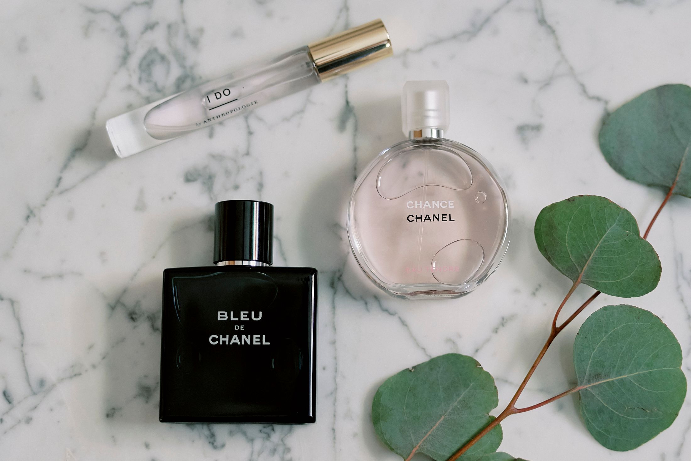 Two Chanel perfumes, Bleu de Chanel and Chance, displayed on marble surface with eucalyptus leaves and a perfume roller.