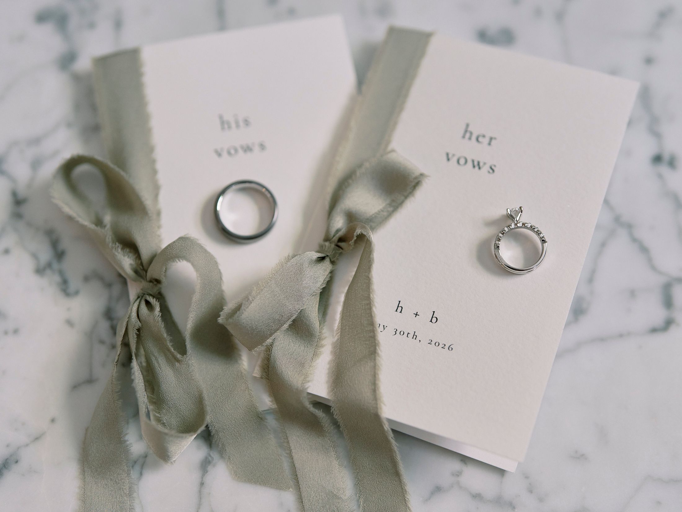 His and her vows booklets with wedding rings and sage green silk ribbons on marble background.