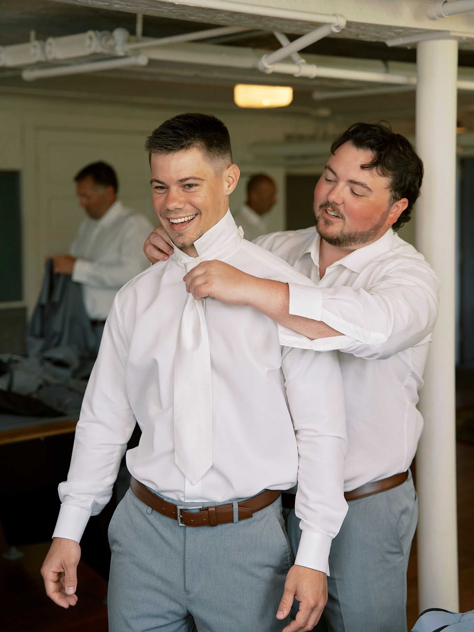Groomsman helping groom fix his white tie while getting ready for wedding ceremony.
