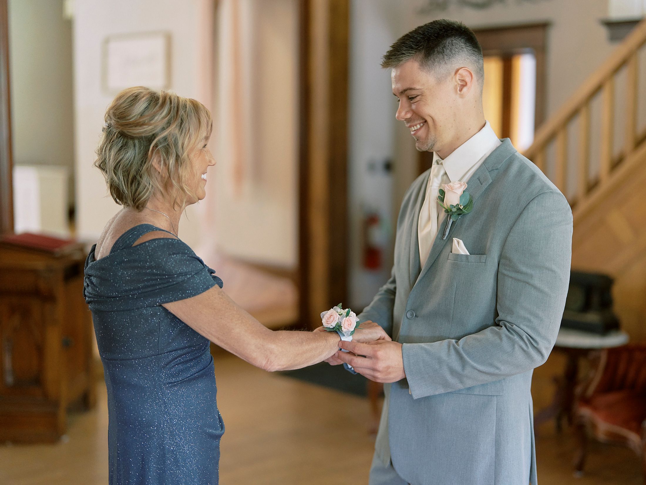 Groom in gray suit smiles as woman pins boutonniere on his lapel during wedding preparations indoors.