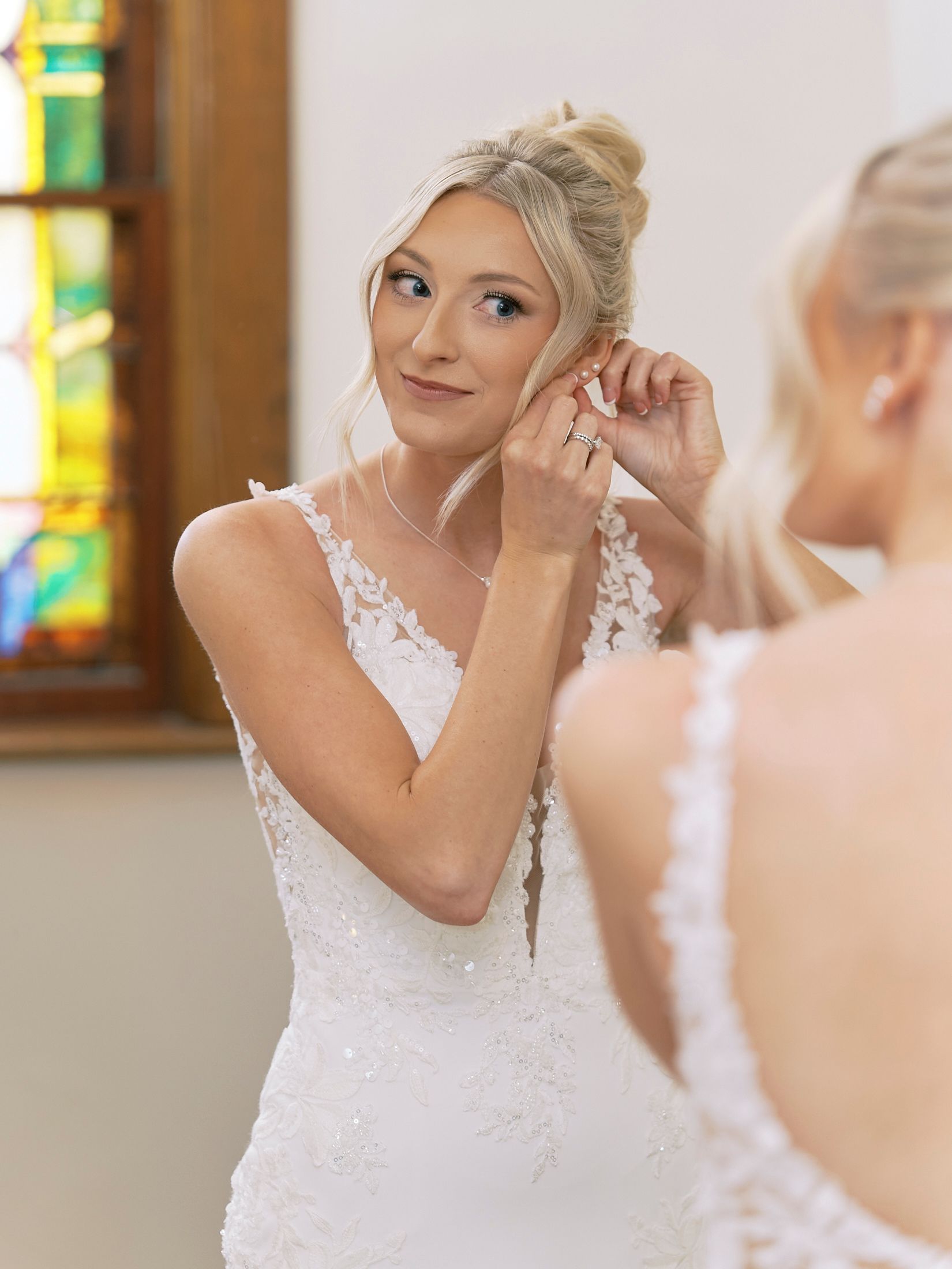 Bride in lace wedding dress putting on earrings while looking in mirror, with stained glass window in background.