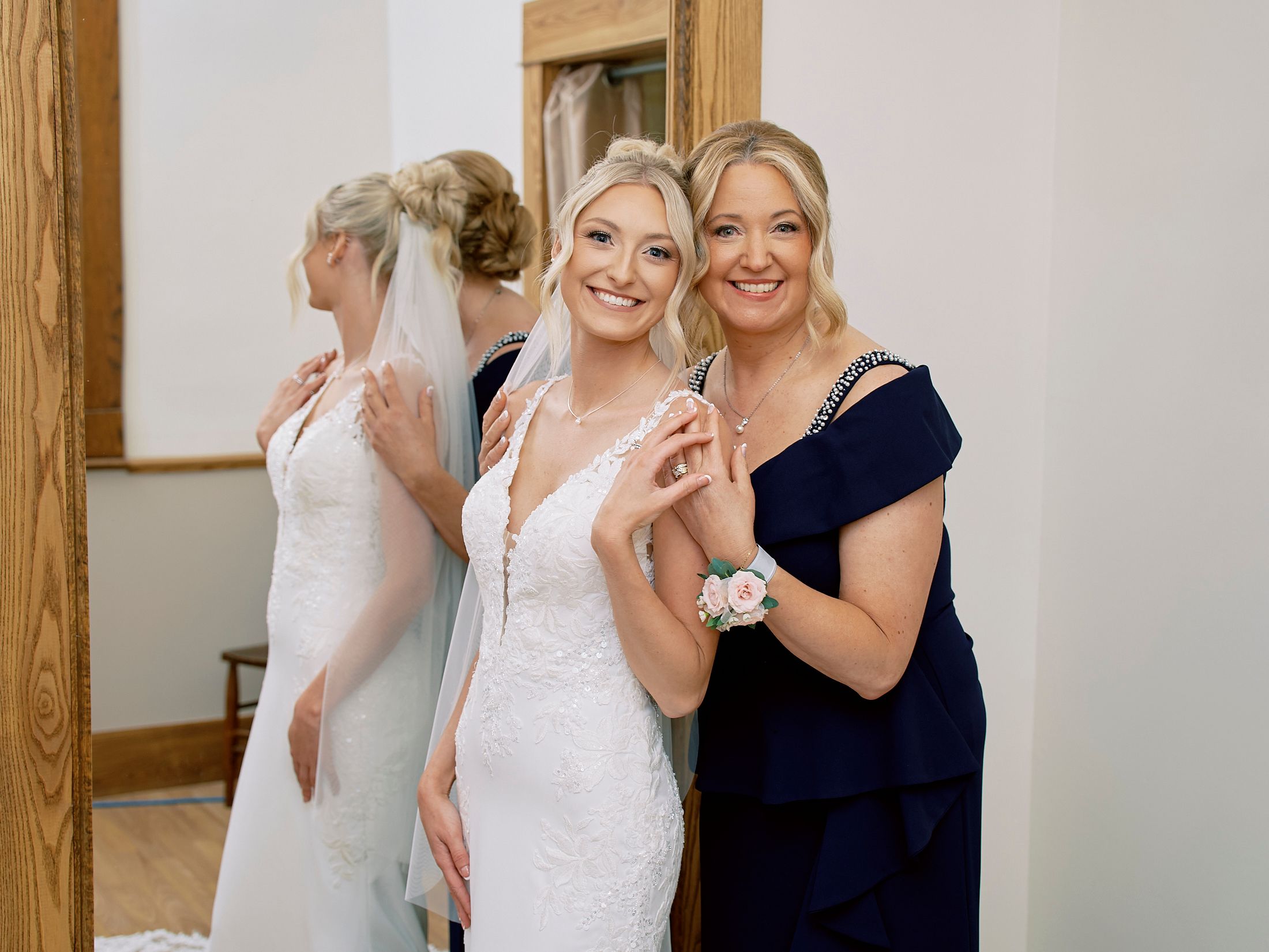Bride in lace wedding gown poses with smiling woman in navy dress during wedding preparations indoors.