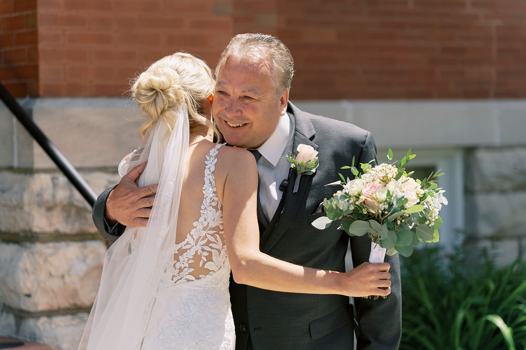 Groom smiling and embracing bride in lace wedding dress outdoors, bride holding white floral bouquet.