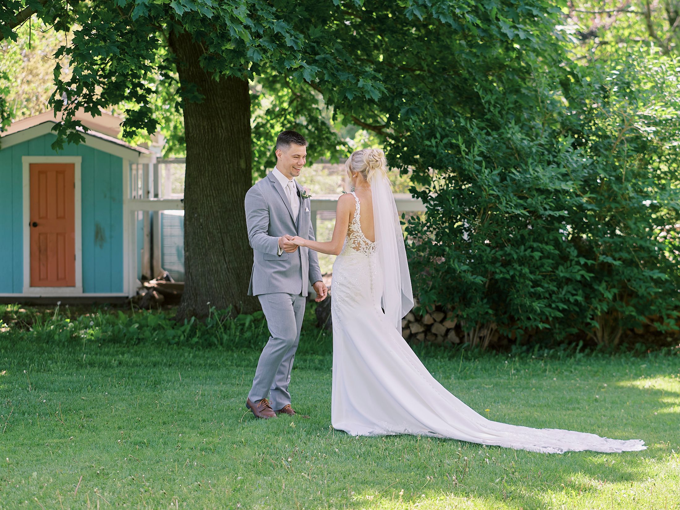 A bride and groom share a tender first look moment on a lush green lawn beside a charming blue garden shed.