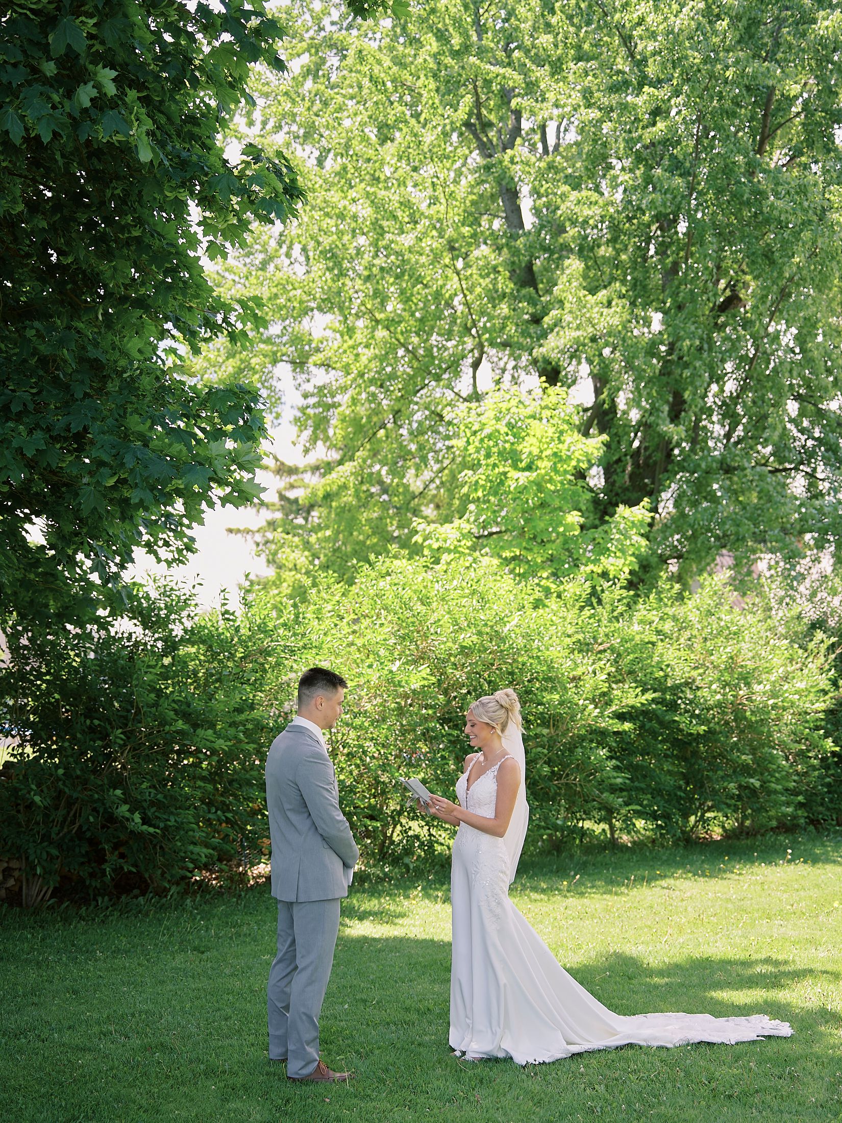 Bride and groom exchange vows outdoors on green lawn surrounded by lush trees on their wedding day.