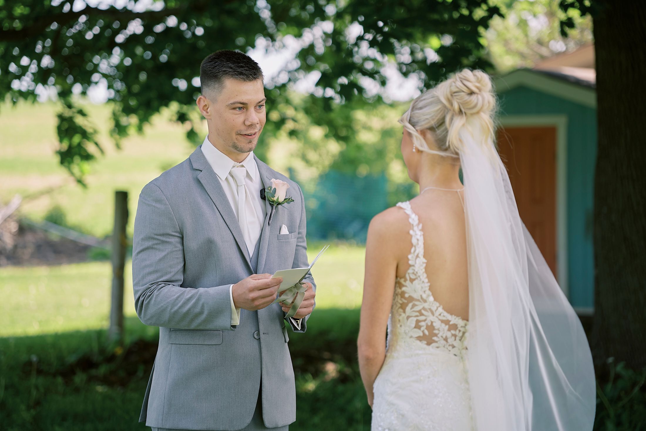 Groom in gray suit reads vows to bride in lace wedding dress during outdoor first look ceremony.