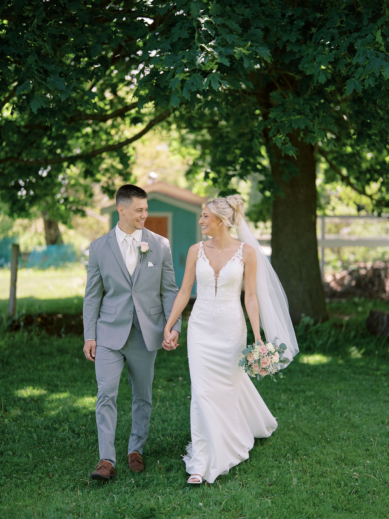 Bride and groom walk hand-in-hand outdoors under lush green trees on their wedding day.