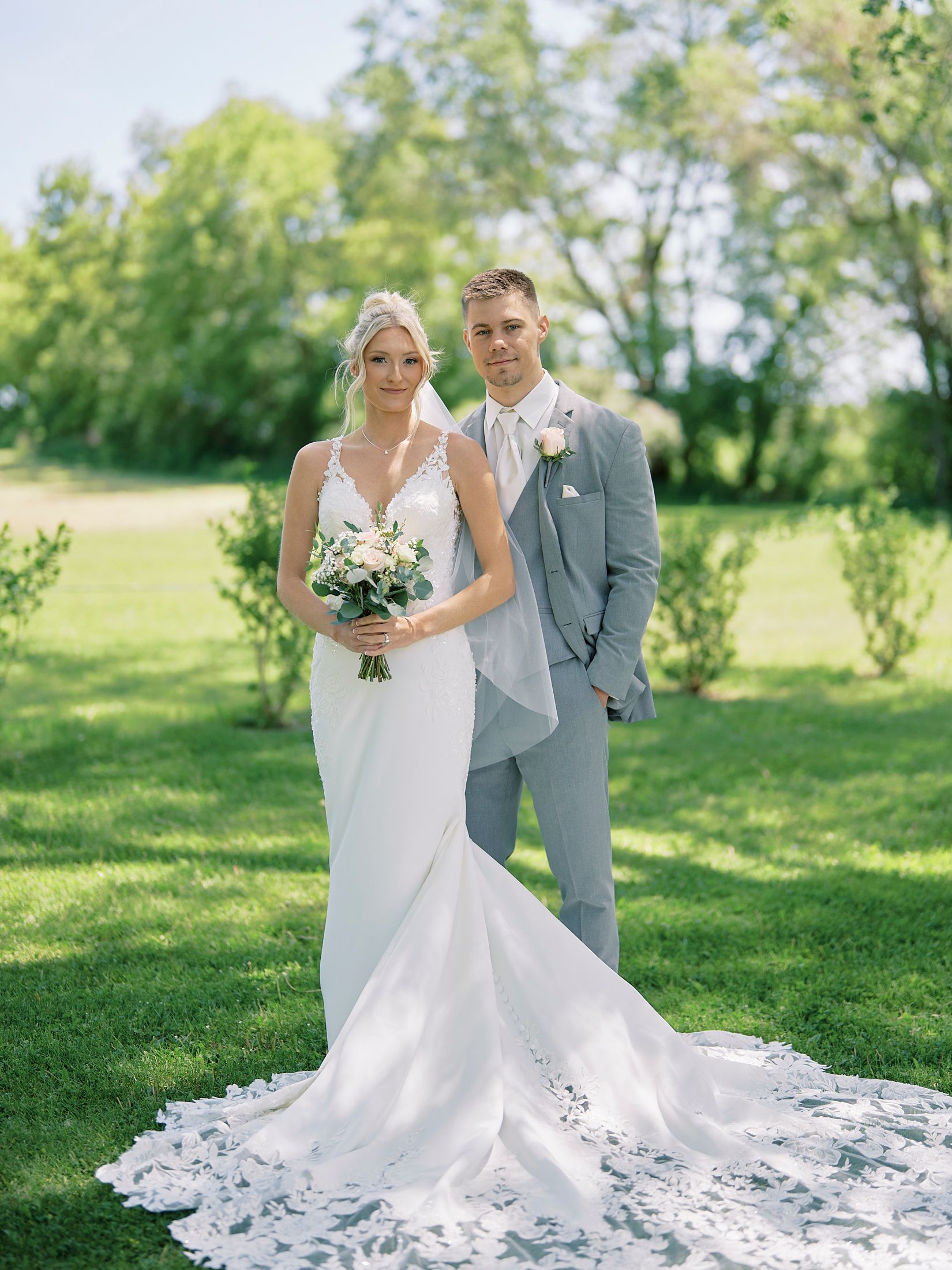 Bride in white gown with lace train holds bouquet beside groom in gray suit on green lawn.