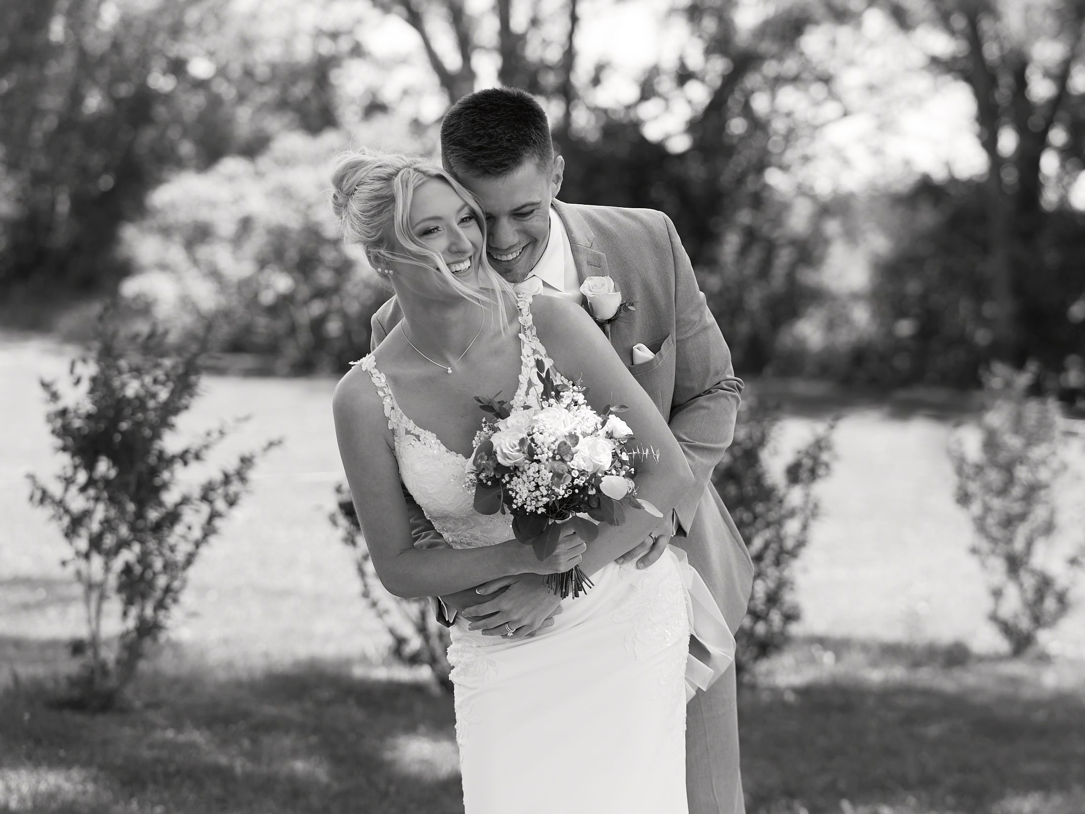 Black and white photo of groom hugging smiling bride holding bouquet in outdoor garden setting.