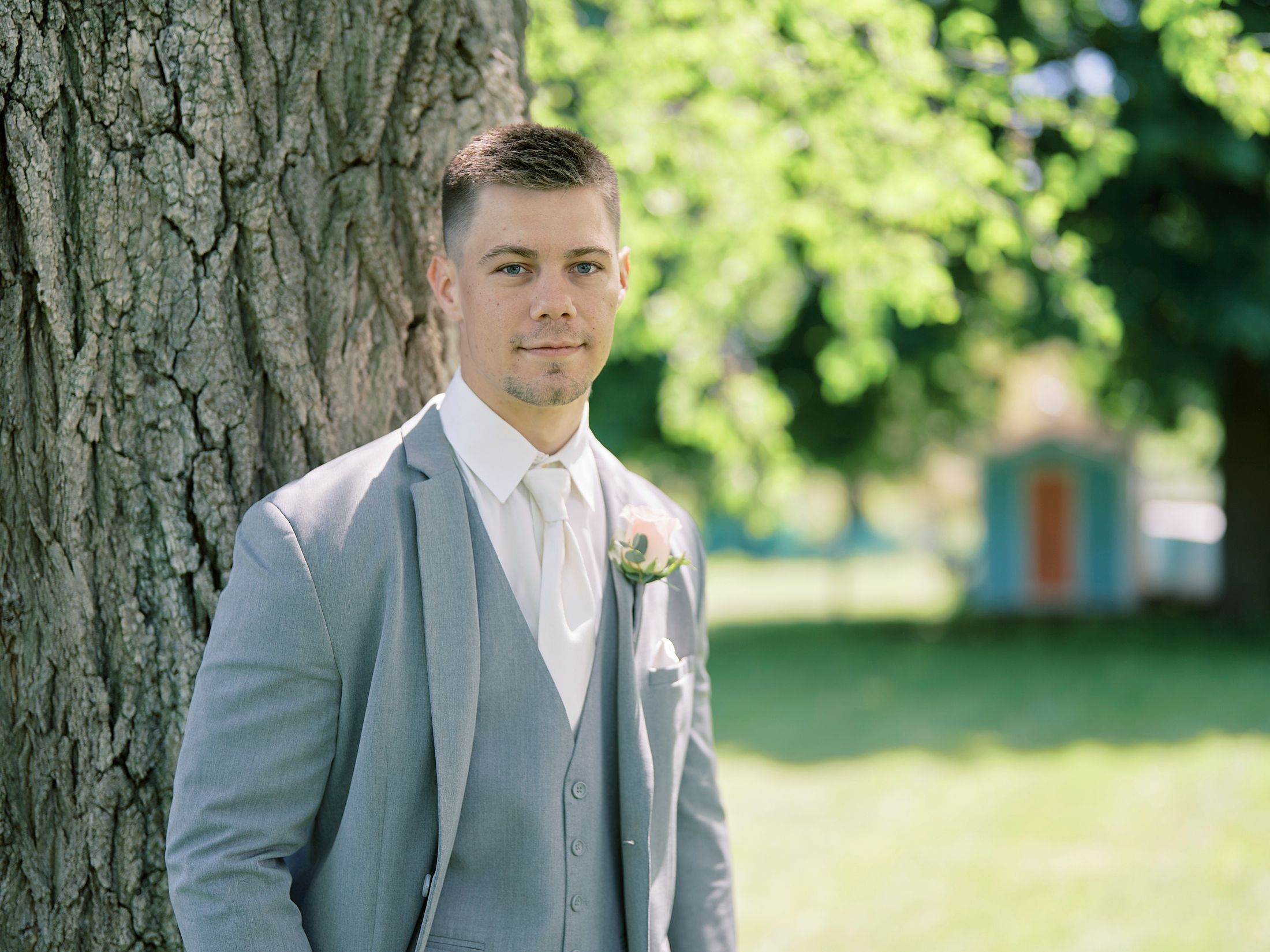 Dapper groom in grey suit with boutonniere leaning against tree outdoors.