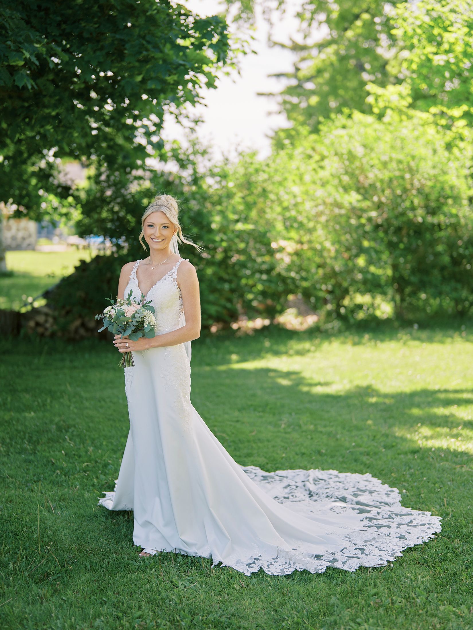 Bride in elegant lace-train wedding gown holding bouquet, standing on green lawn surrounded by lush trees.