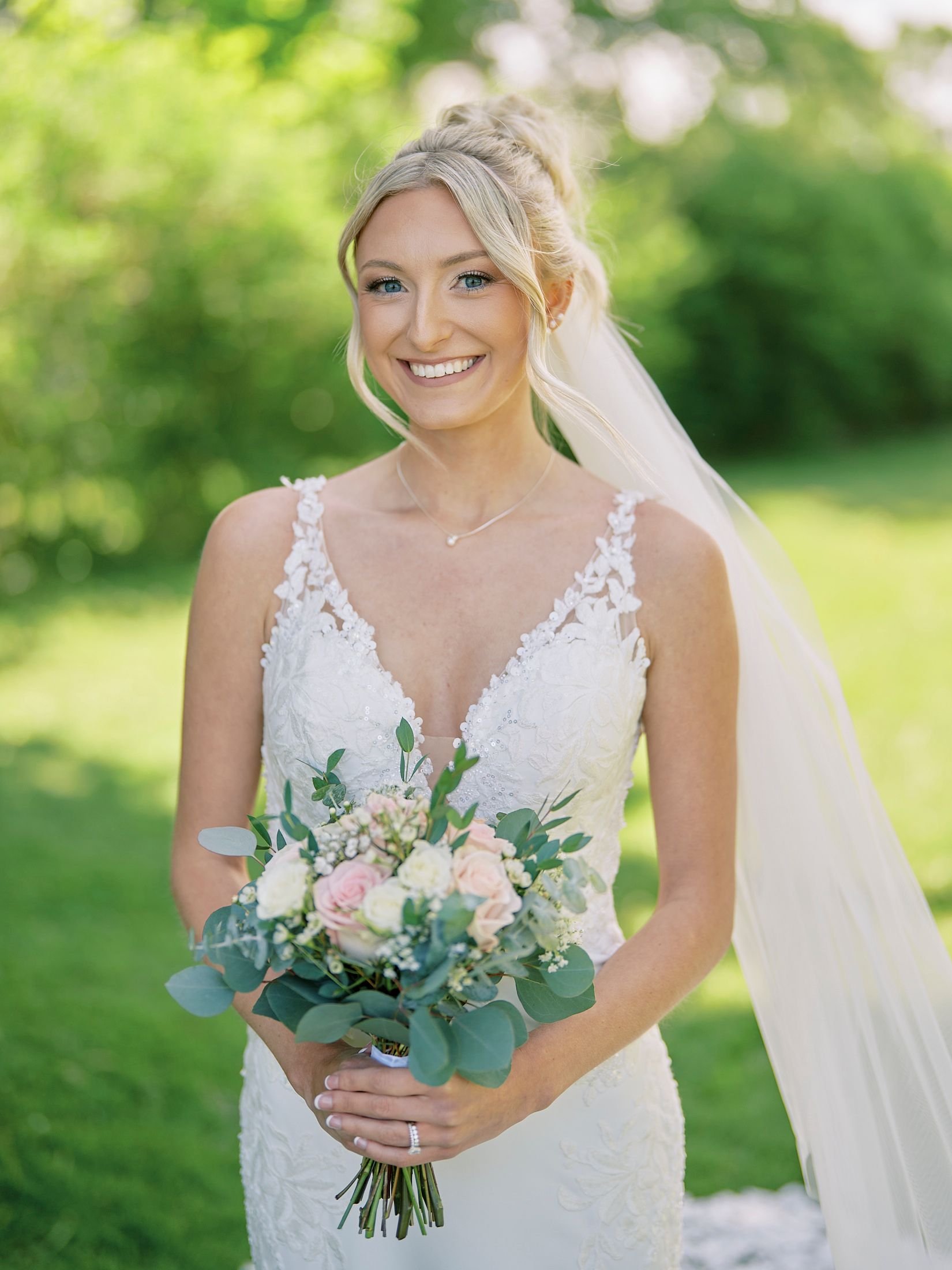 Smiling bride in lace V-neck wedding gown holding blush pink rose bouquet with eucalyptus outdoors.