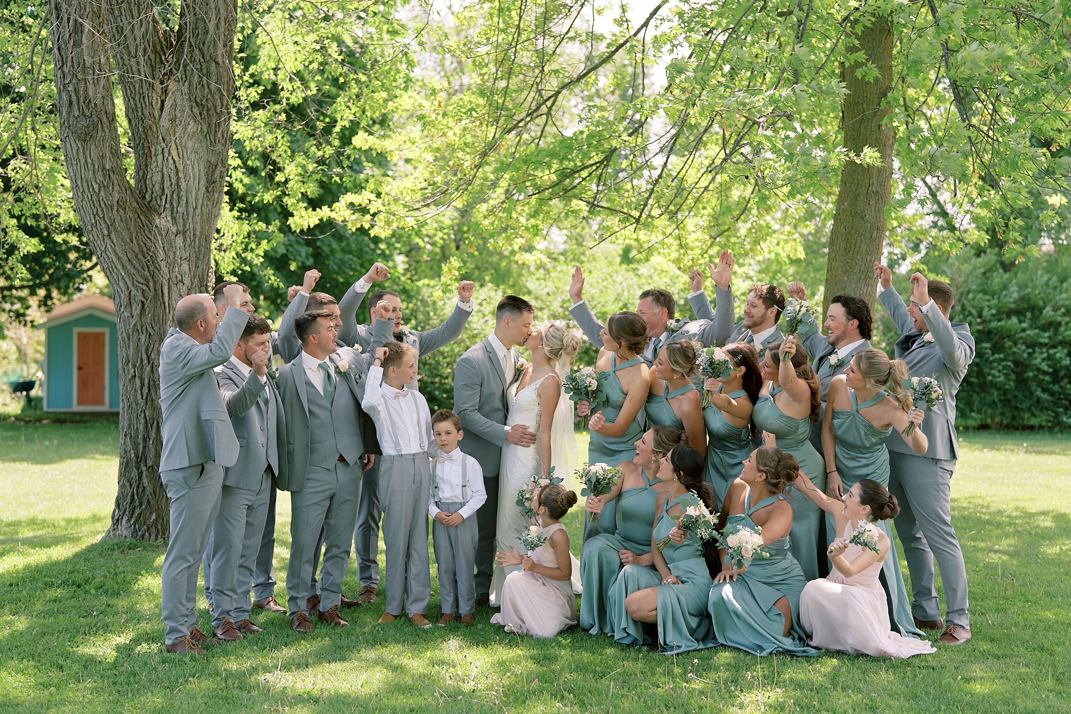 Wedding party celebrating outdoors under trees in sage green and grey attire.