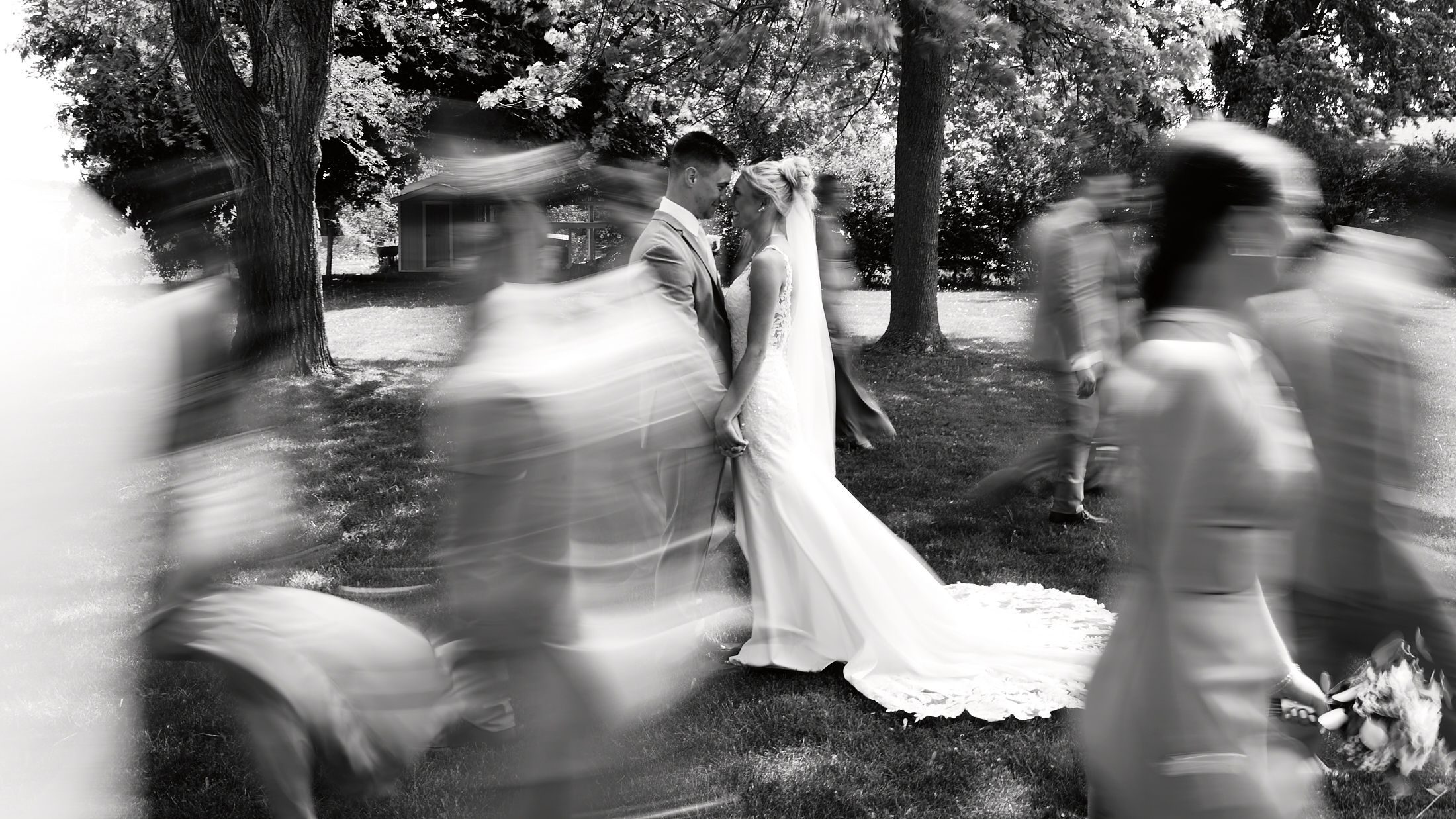 Artistic black and white blurred motion photo of bride and groom surrounded by wedding party outdoors.