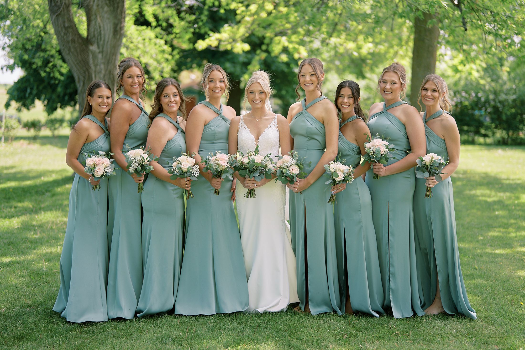 Bride in white gown poses with eight bridesmaids in sage green dresses holding bouquets.