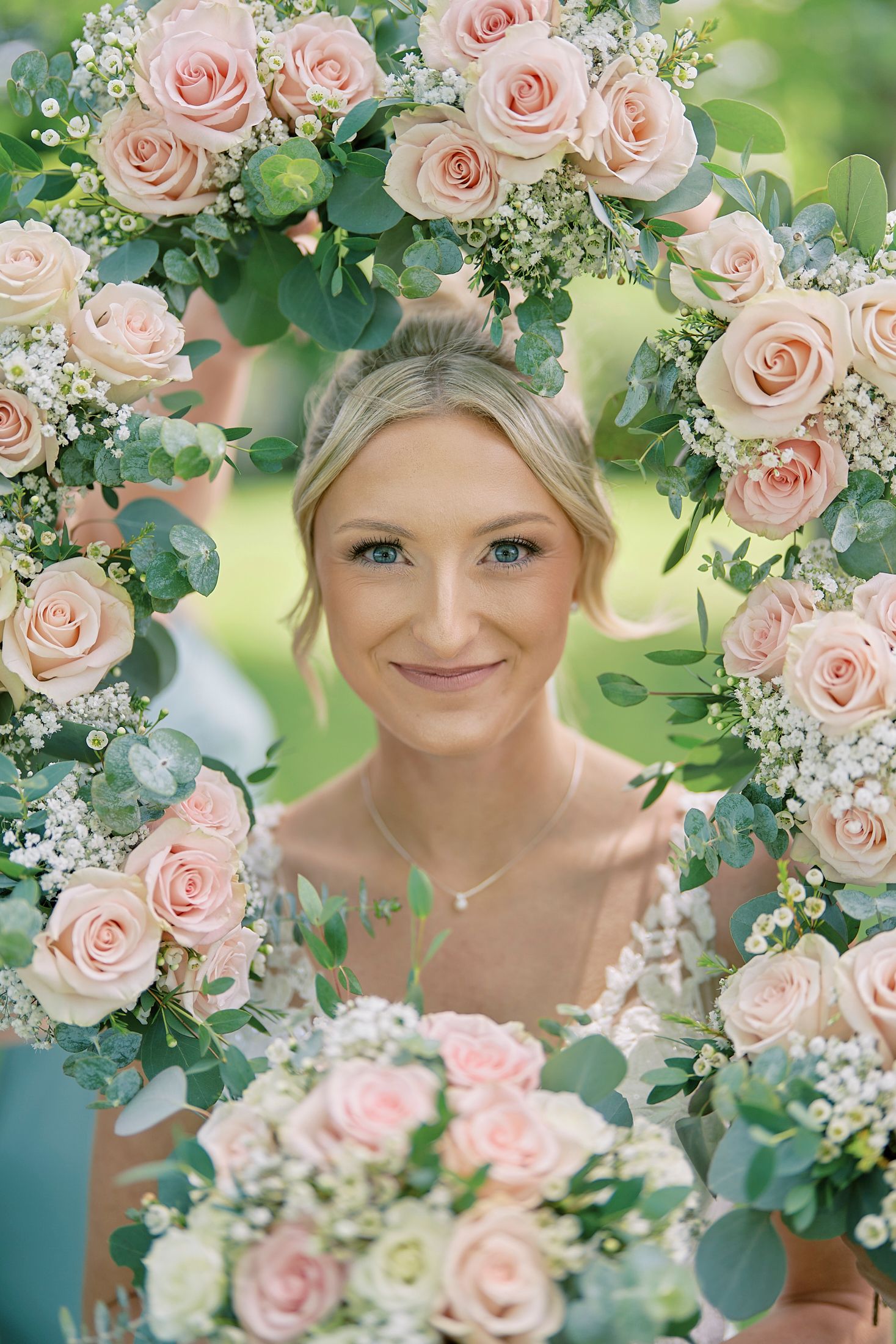 Bride smiling through a floral frame of blush pink roses and eucalyptus, holding a matching bouquet.