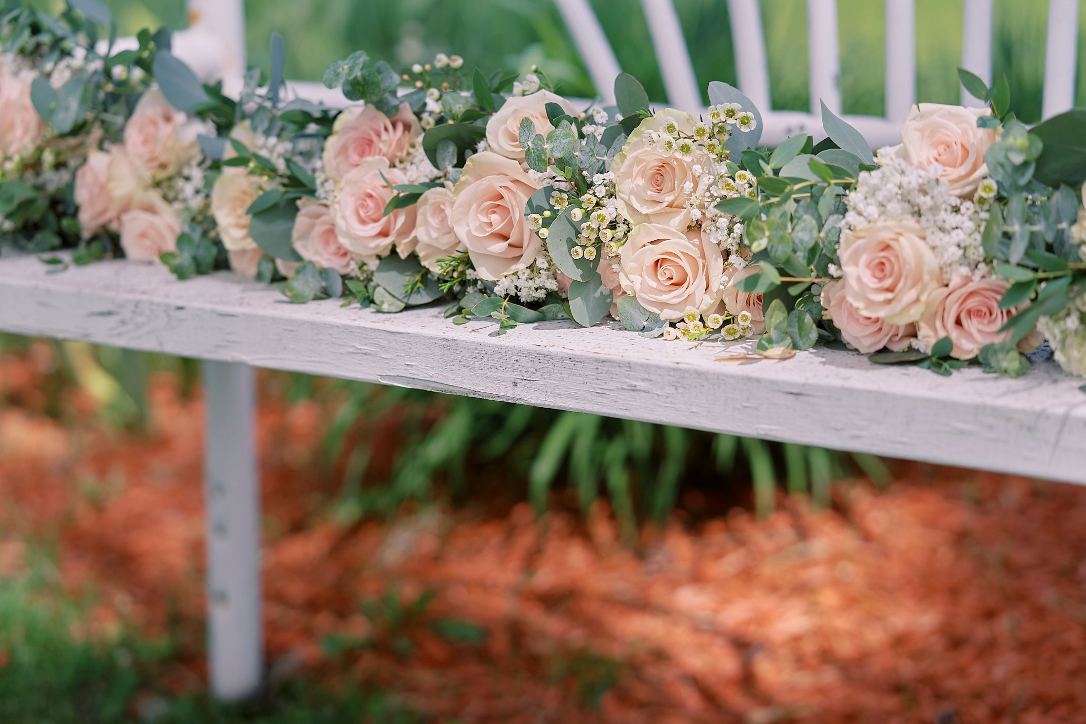 Blush pink roses and eucalyptus bridesmaid bouquets arranged on a rustic white wooden bench.