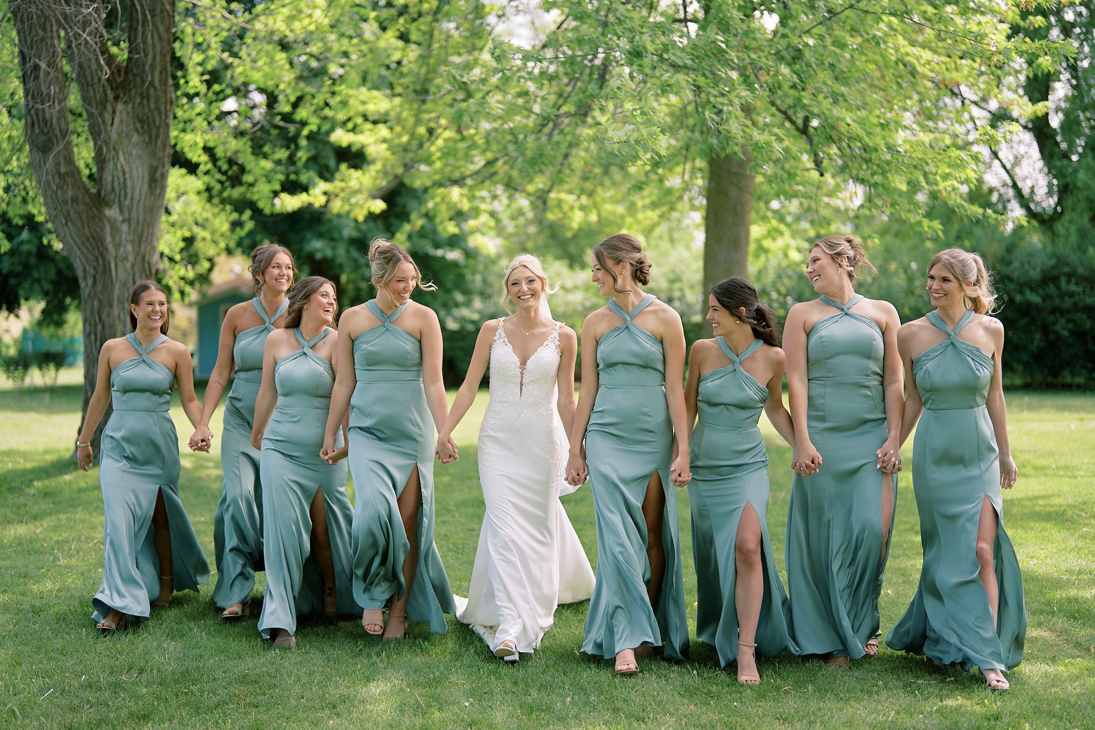 Bride in white gown walks with bridesmaids in sage green dresses outdoors on lush green lawn.