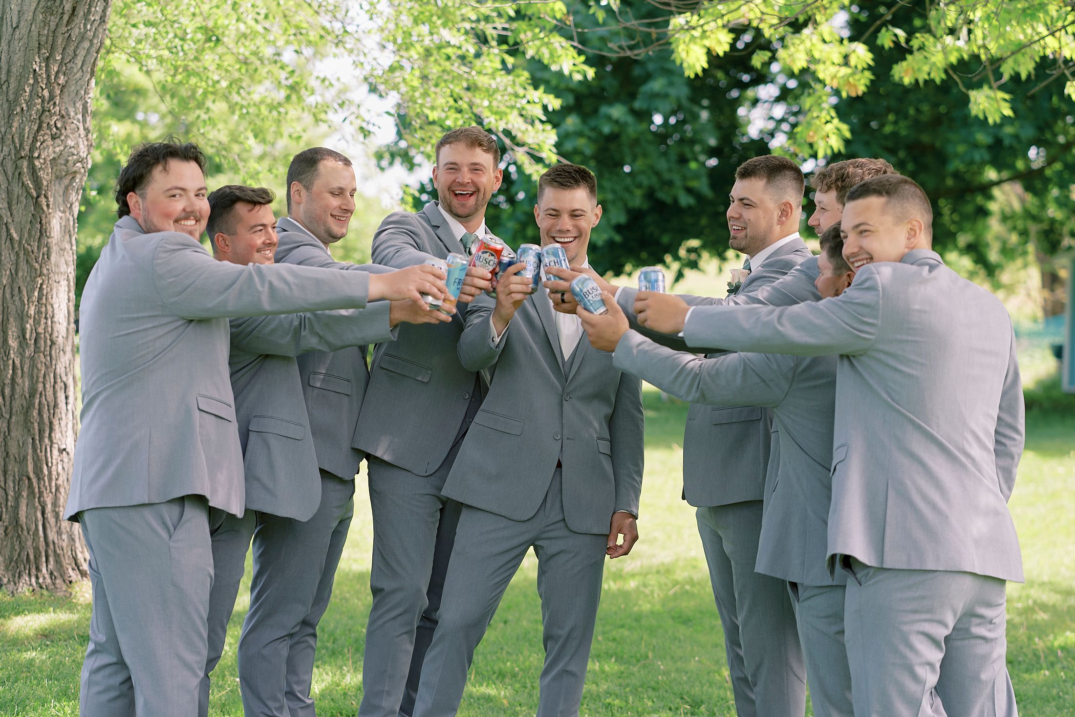 Groomsmen in gray suits toasting with drinks outdoors under green trees at a wedding celebration.