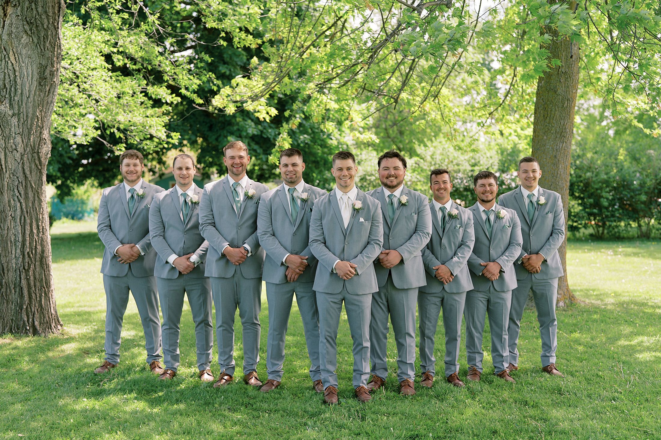 Groom and groomsmen posing in matching light gray suits with boutonnieres under a large tree on a sunny day.