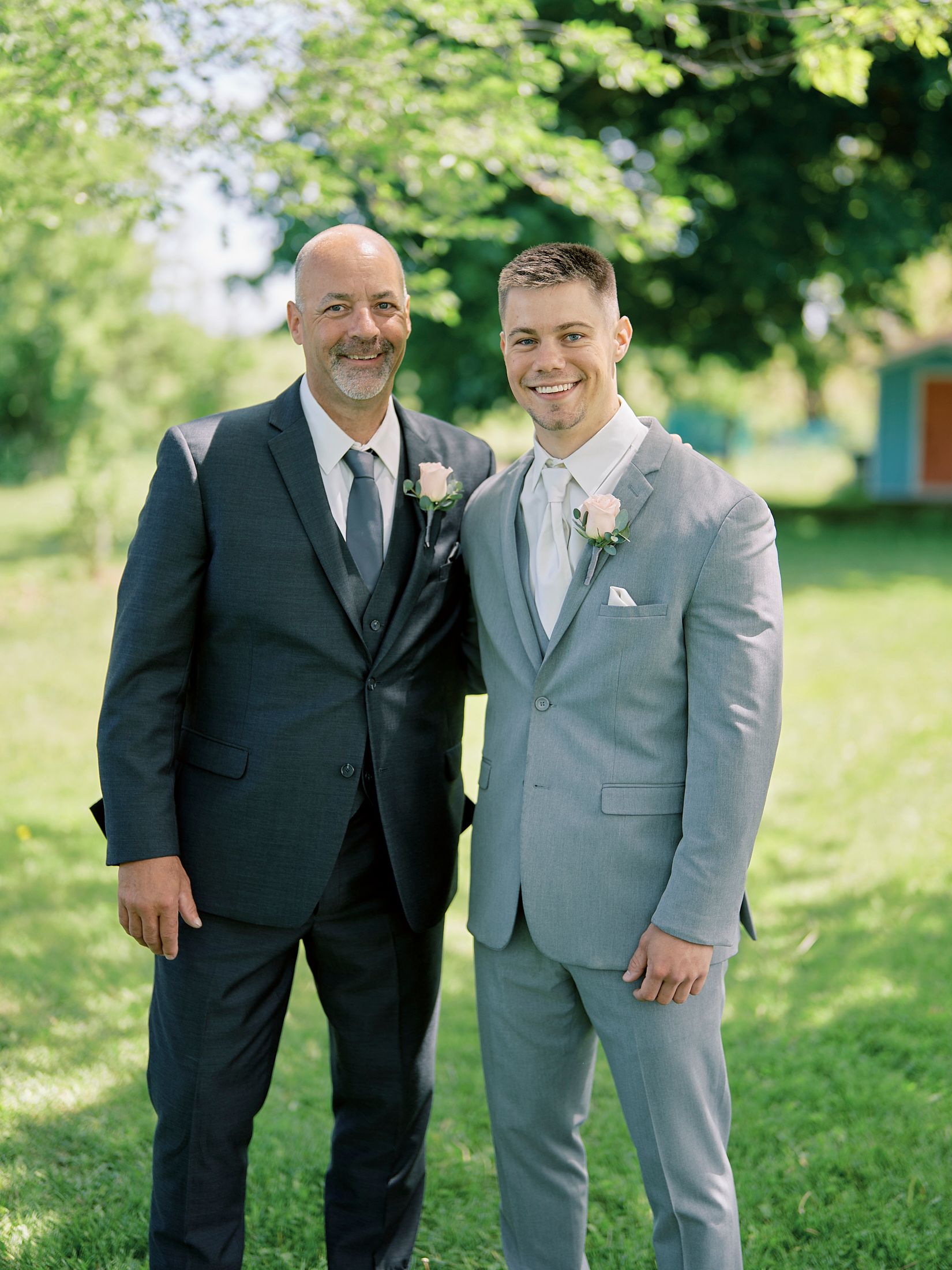 Groom in light gray suit poses with man in dark navy suit at outdoor wedding venue.