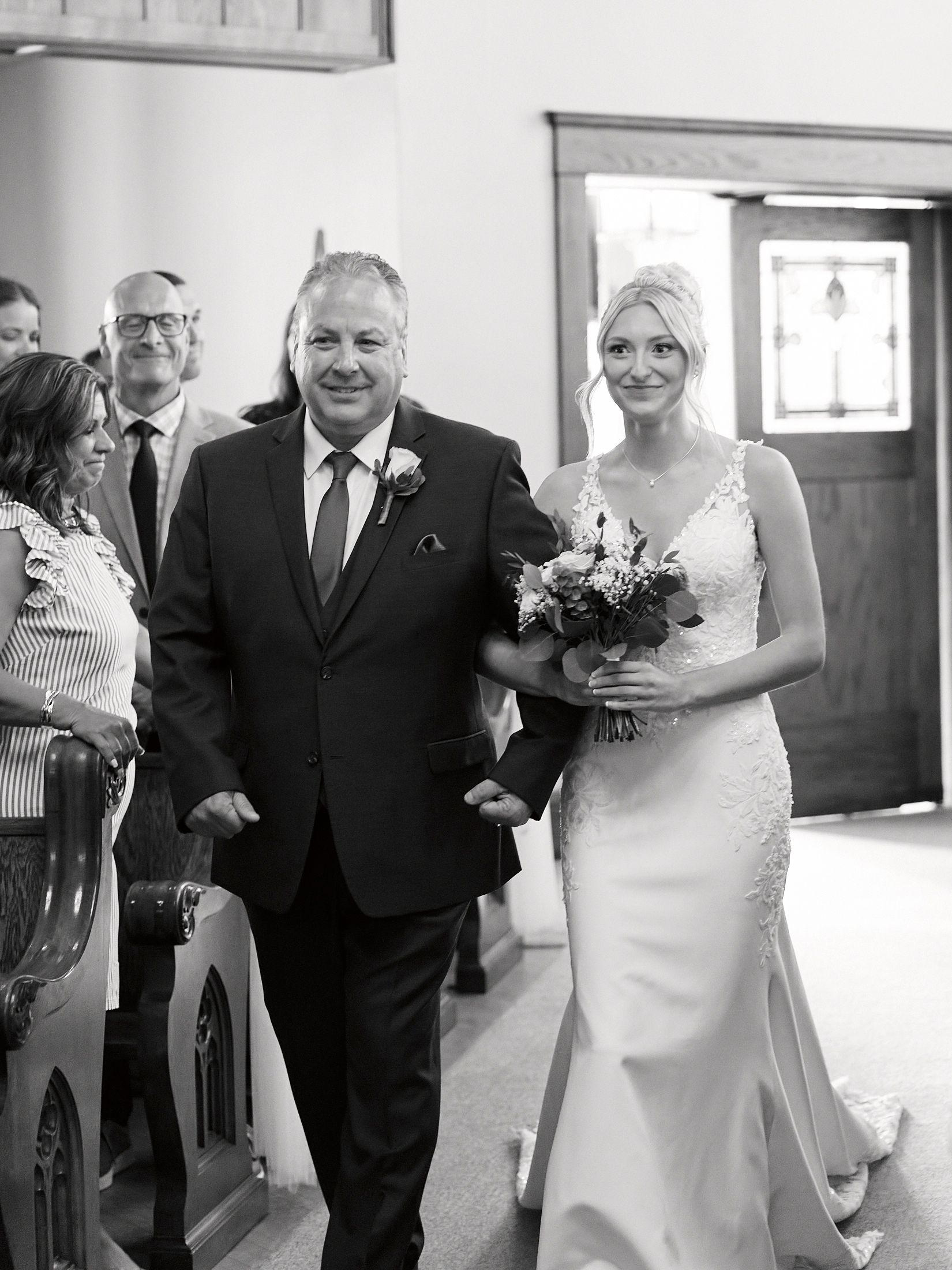 Bride walking down the aisle with her father in a black and white wedding ceremony photo.