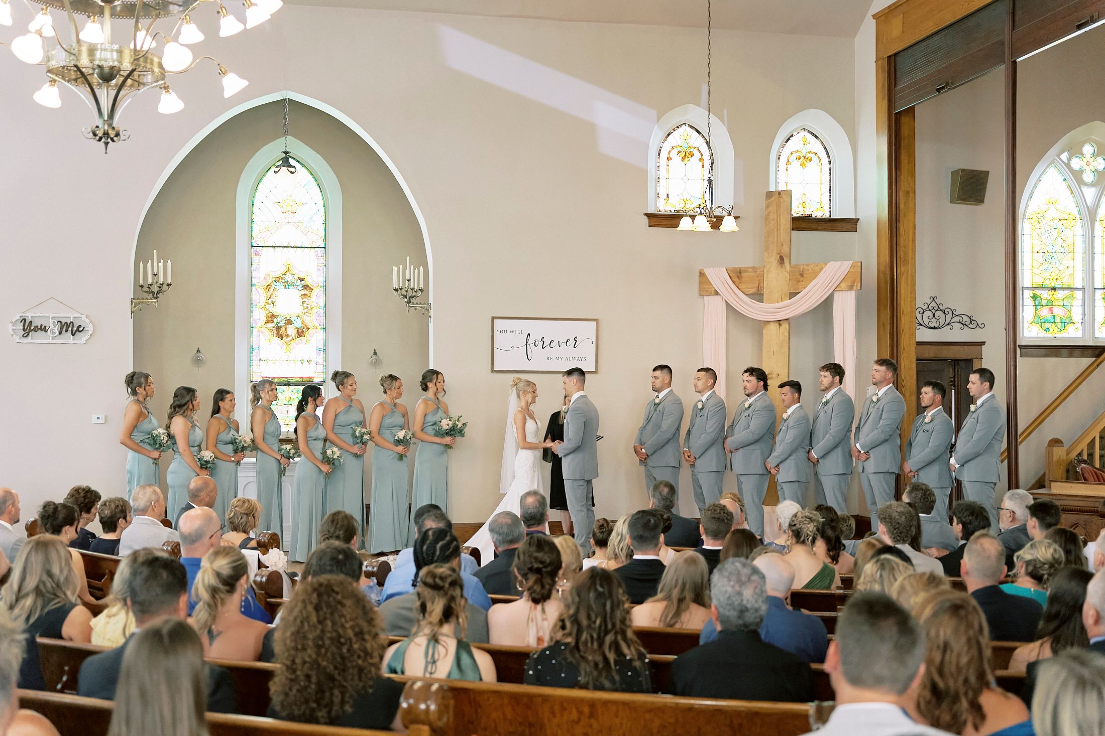 Wedding ceremony in elegant church with large bridal party lined up before guests seated in wooden pews.