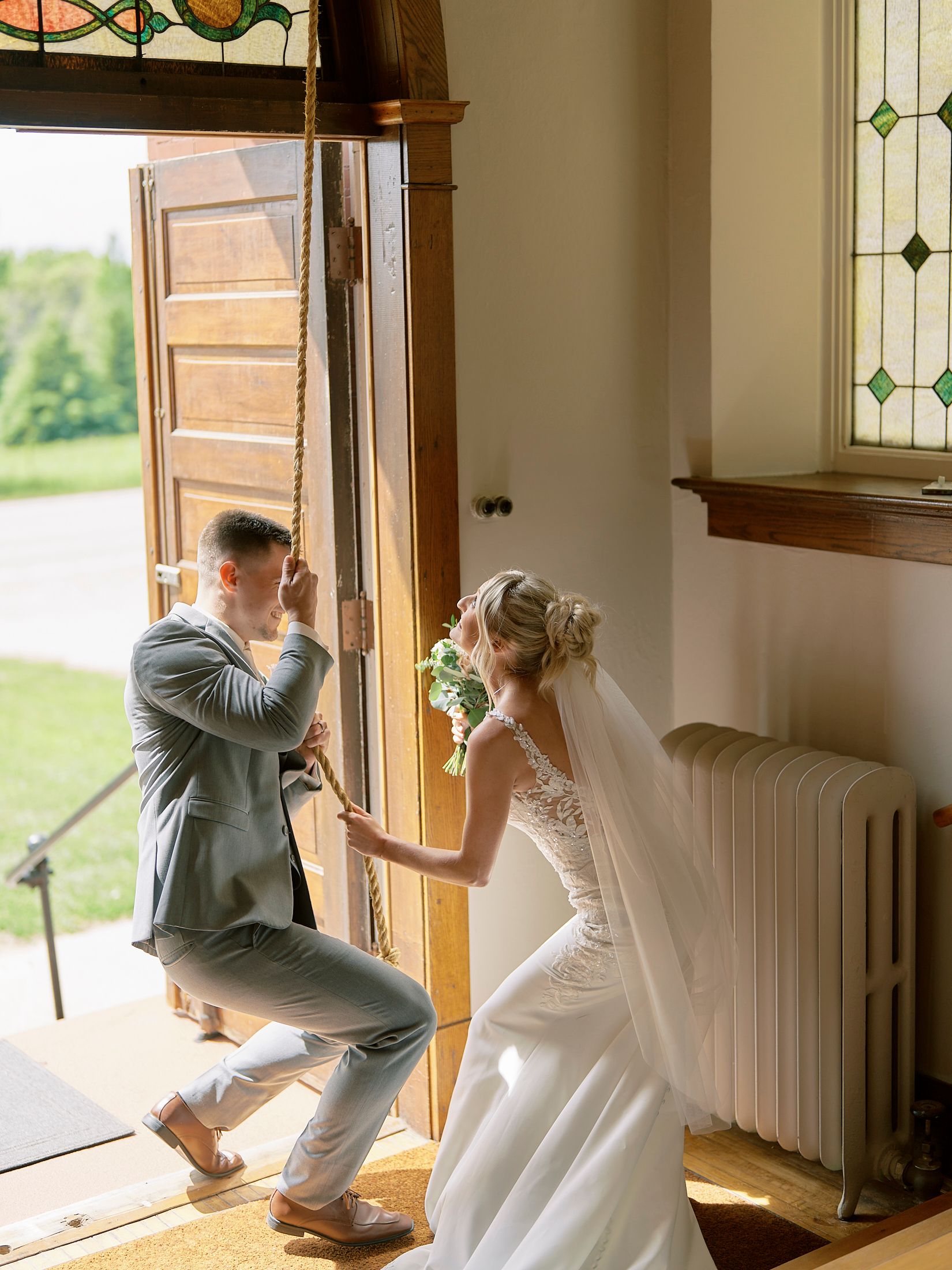 Bride and groom pulling open large wooden church doors with stained glass windows on their wedding day.