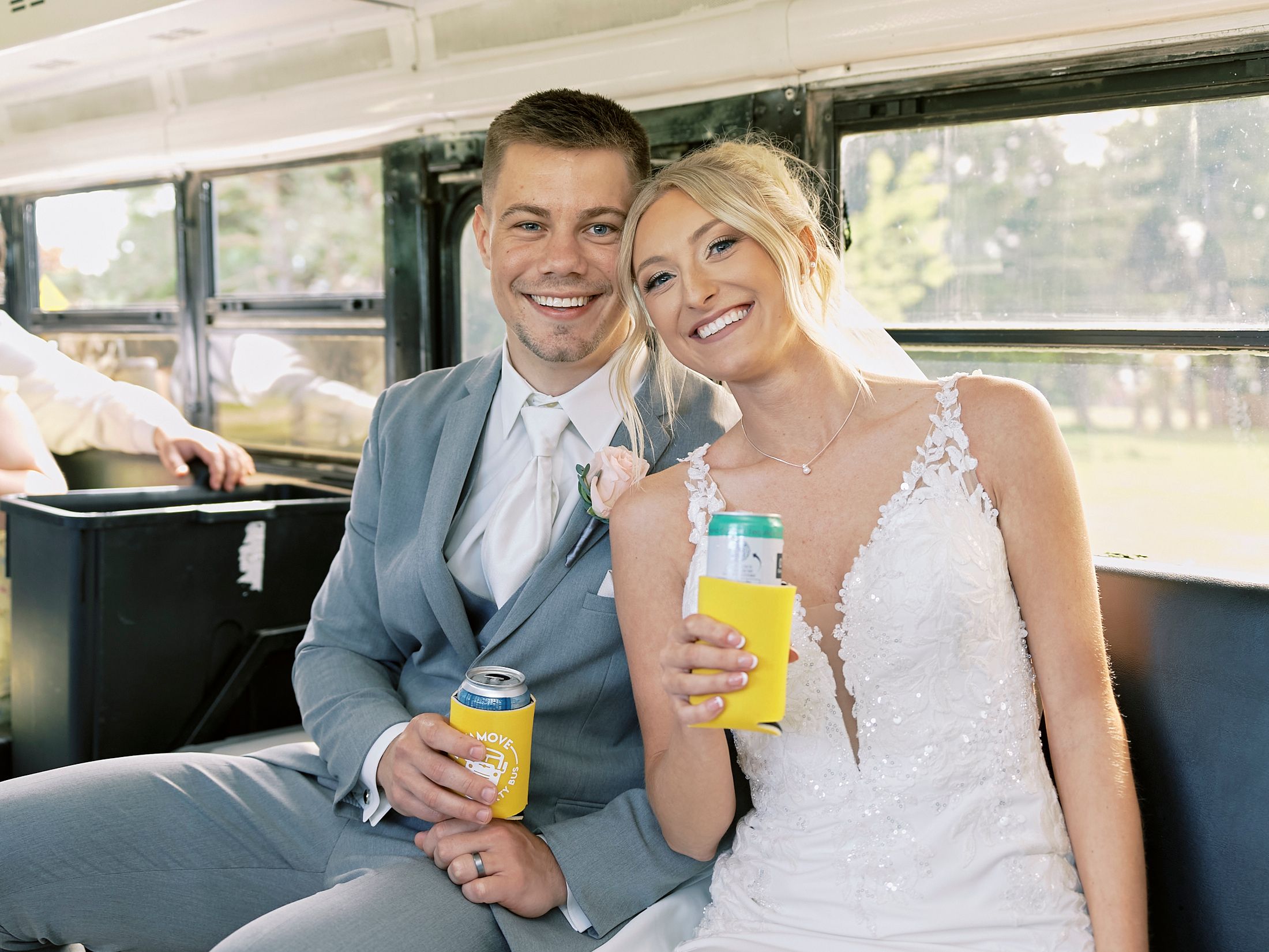 Smiling bride and groom hold canned drinks while seated together on a party bus after wedding.